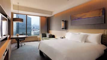 Una habitación de hotel moderna con una cama grande, un escritorio, una silla y vistas a un paisaje urbano a través de amplias ventanas.