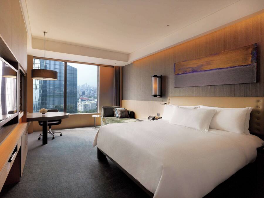 Una habitación de hotel moderna con una cama grande, un escritorio, una silla y vistas a un paisaje urbano a través de amplias ventanas.