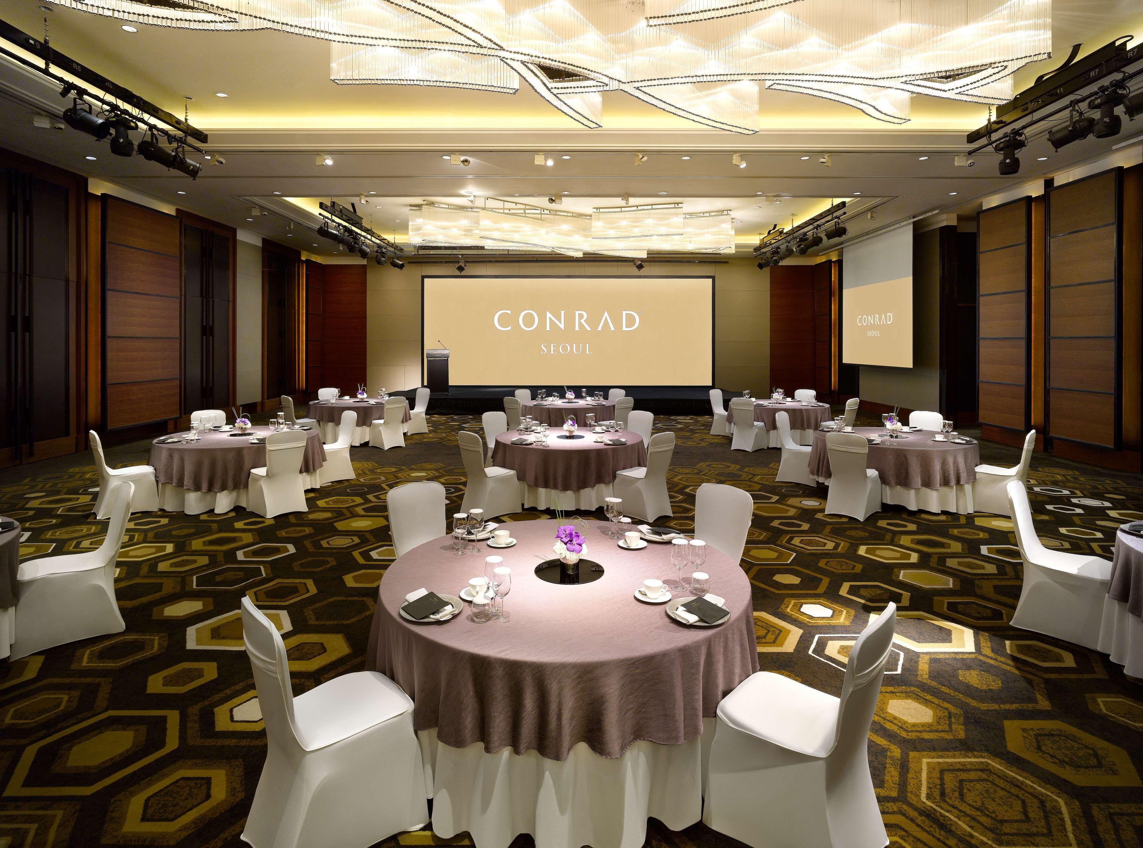 A conference room at Conrad Seoul hotel with multiple round tables, white chairs, and a large screen displaying the hotel name.