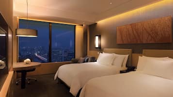 A hotel room with two beds, a desk with a chair, a large window with a city view, and a wall-mounted artwork.