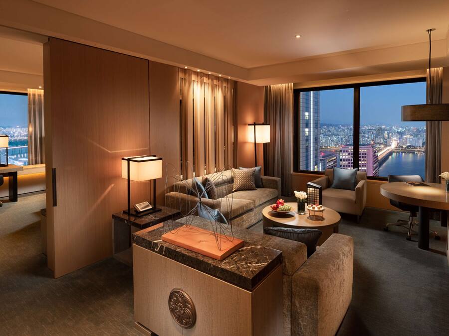 Habitación de hotel moderna con un amplio ventanal que ofrece vistas a la ciudad, una zona de estar confortable con sofá y sillones, una pequeña mesa de comedor y un escritorio con lámpara.