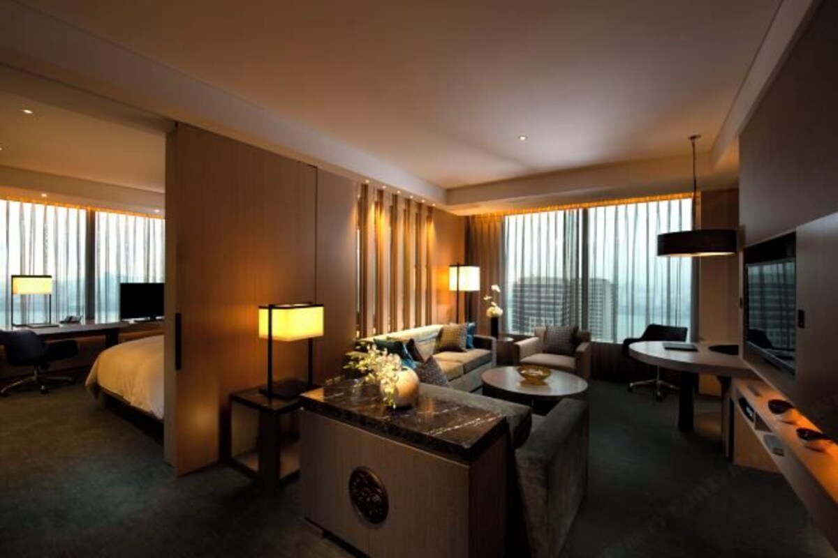 A modern hotel room with a bed, a desk, a sofa, and a small dining table. There is a large window with curtains, a TV, and a lamp.