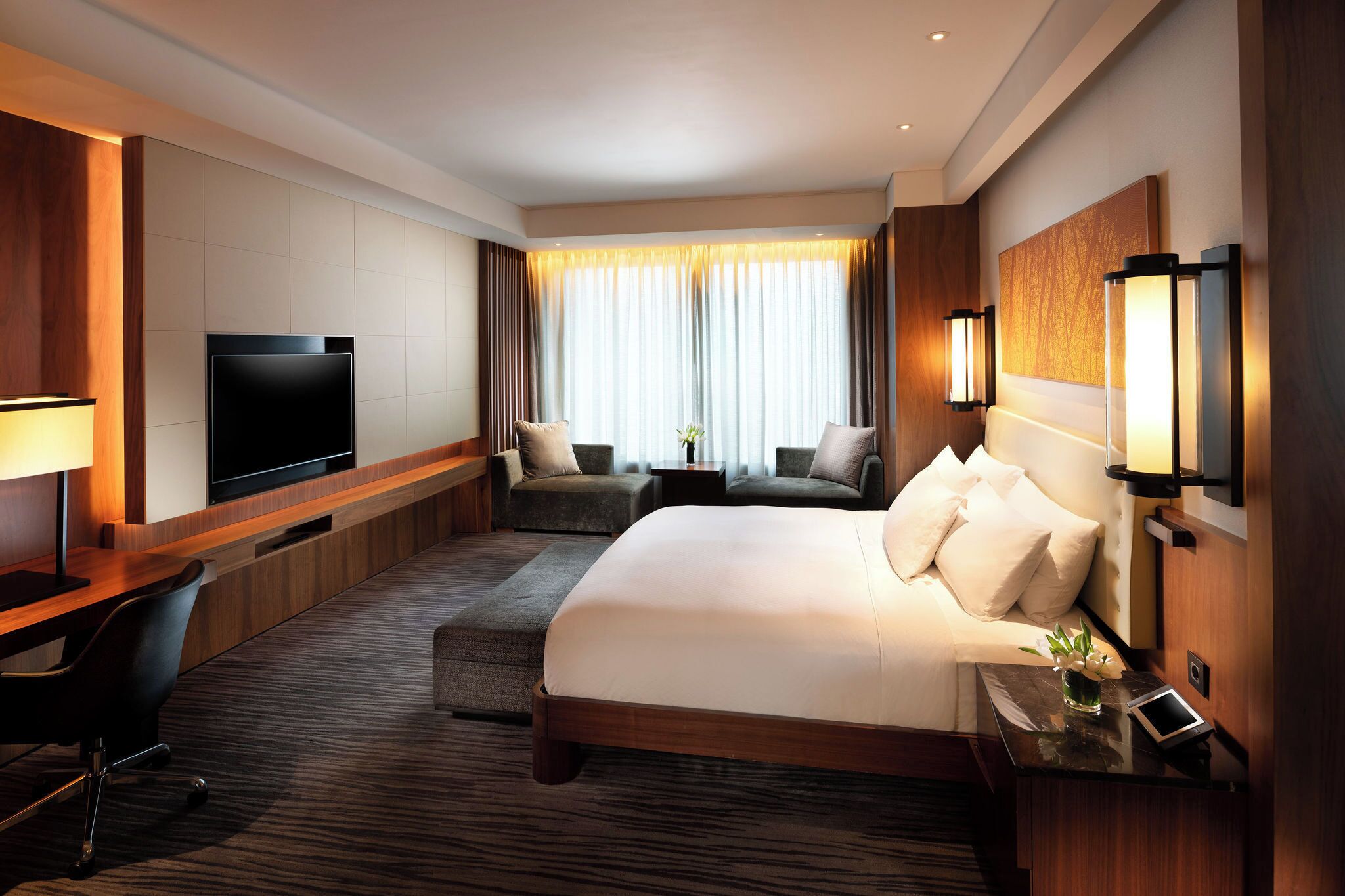 A hotel room with a large bed, a desk with a chair, a flat-screen TV, a sofa, and a small table with a lamp.