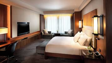 A hotel room with a large bed, a desk with a chair, a flat-screen TV, a sofa, and a small table with a lamp.