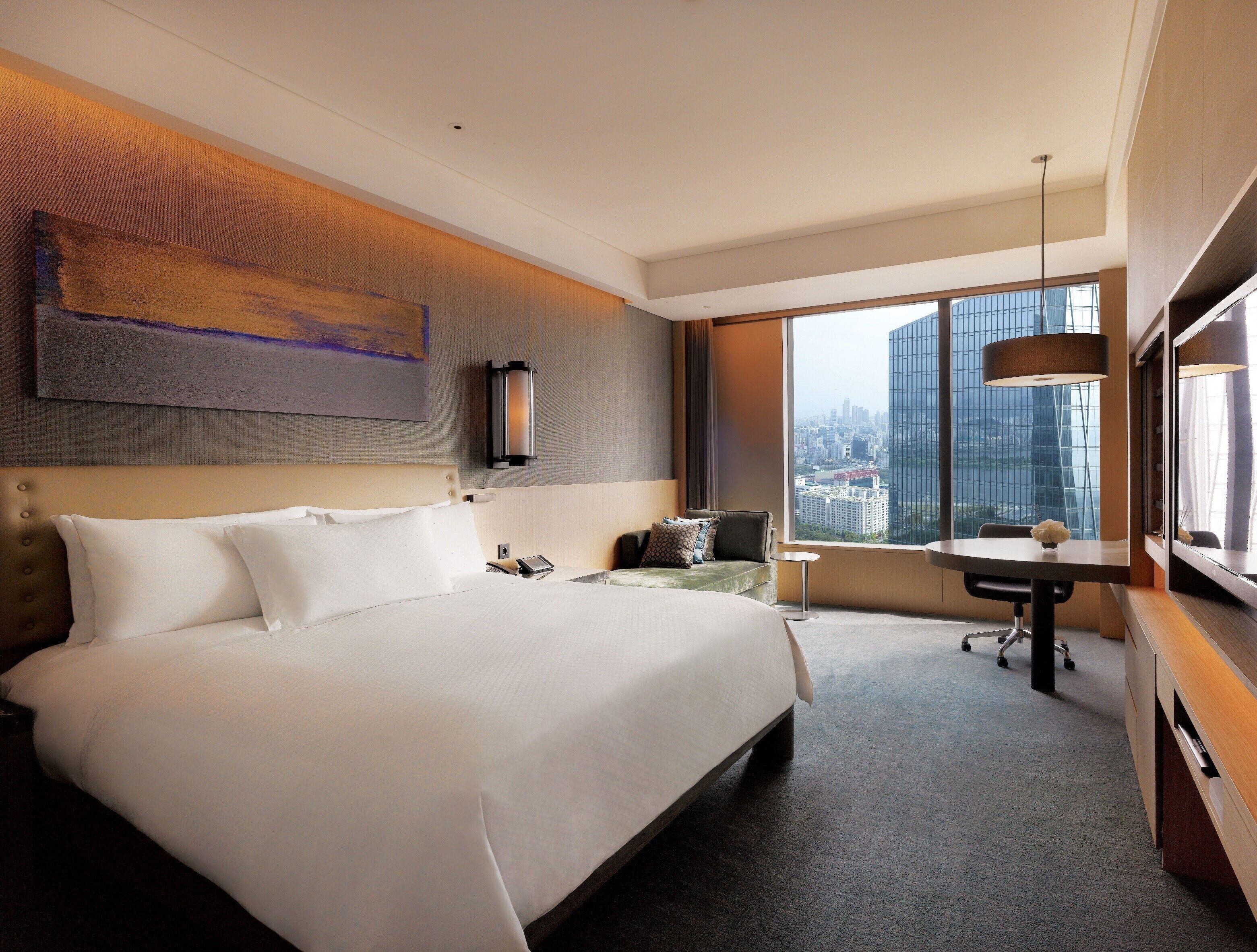 A modern hotel room with a large bed, a desk, and a view of the cityscape.