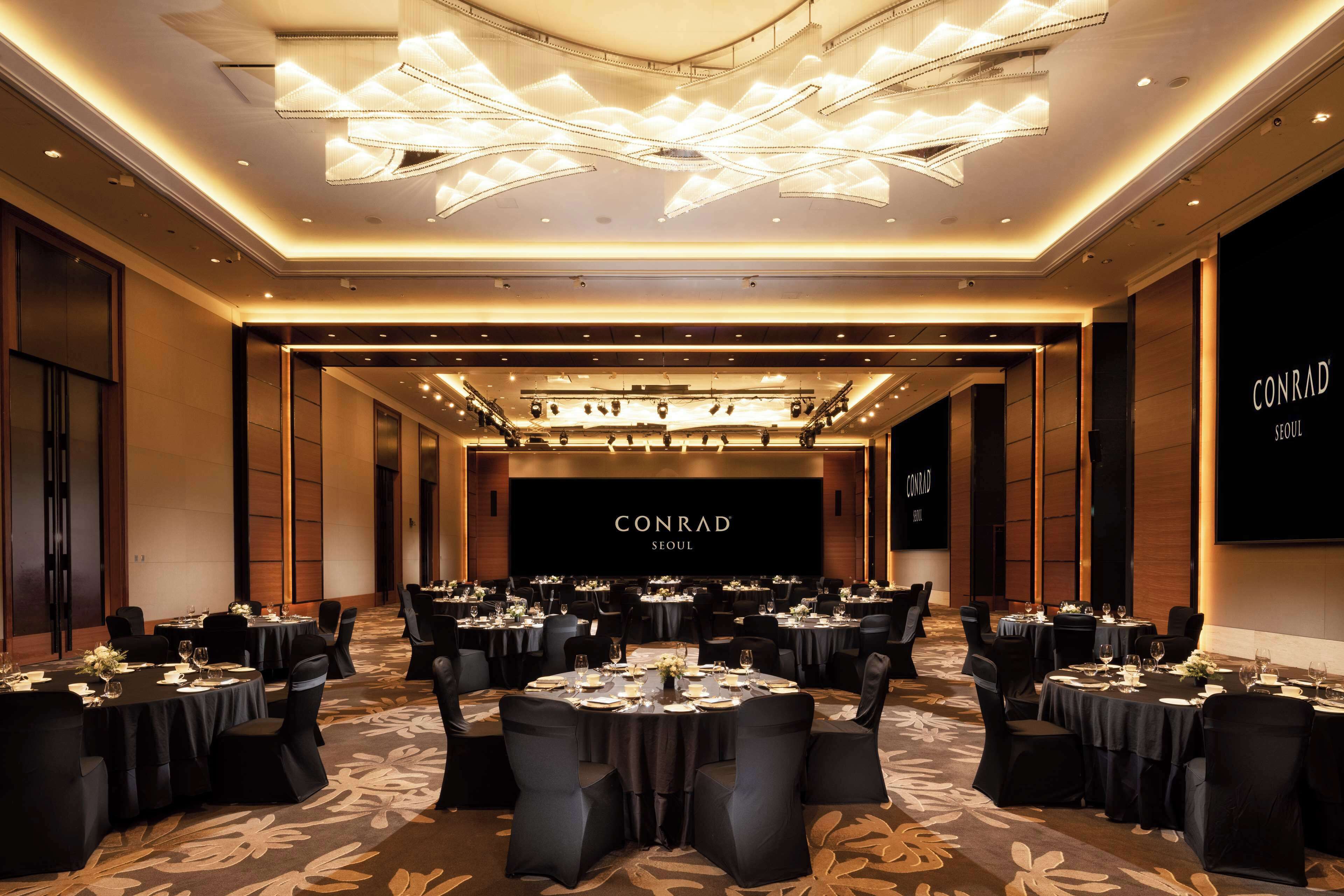 Una amplia sala de conferencias con mesas redondas, sillas negras y una pantalla grande que muestra "CONRAD SEOUL".