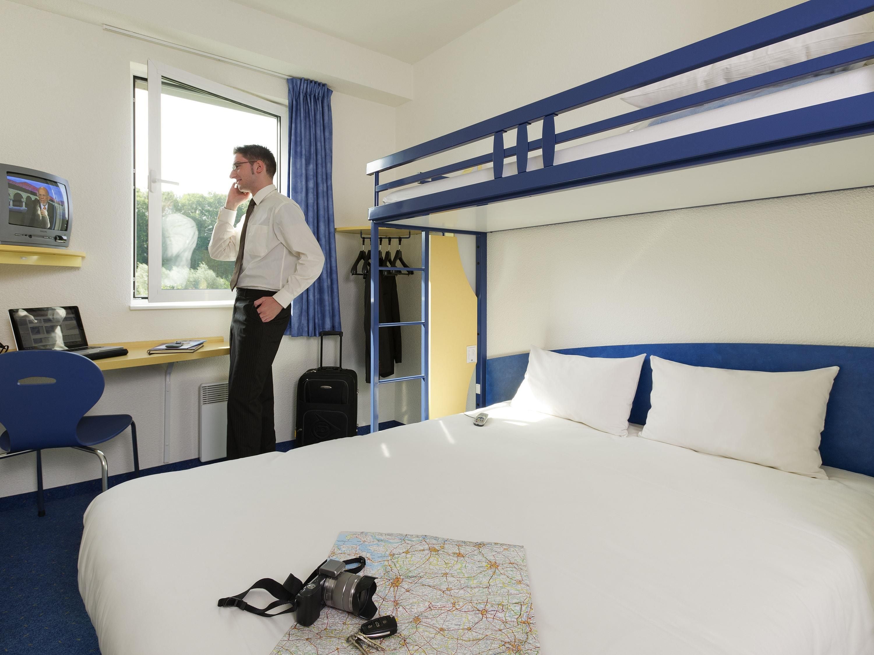 Ein Hotelzimmer mit einem Etagenbett, einem Schreibtisch mit Computer, einem Fernseher und einem Fenster mit Vorhängen.
