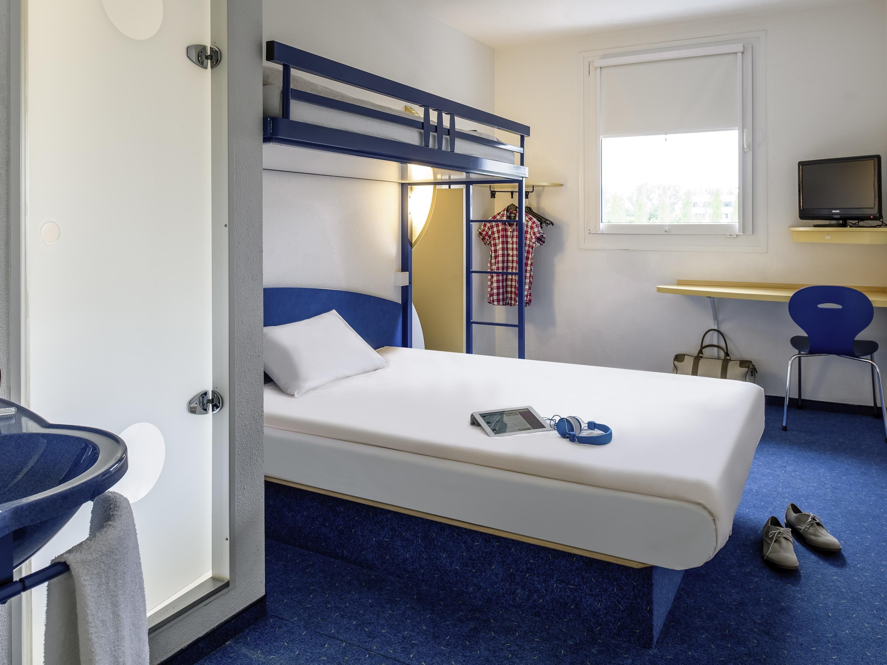 A hotel room with a bunk bed, a desk with a computer, a chair, a TV, and a pair of shoes on the floor.