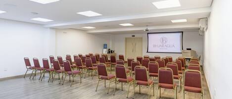 Un salón de conferencias con filas de sillas, una pantalla de proyector mostrando 'BHJARAGUÁ HOTEL' y un dispensador de agua.