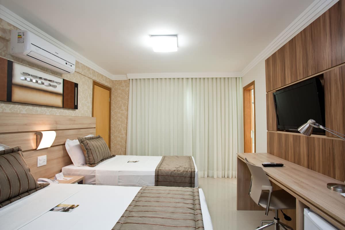 A hotel room with two beds, a desk, a TV, and an air conditioning unit.