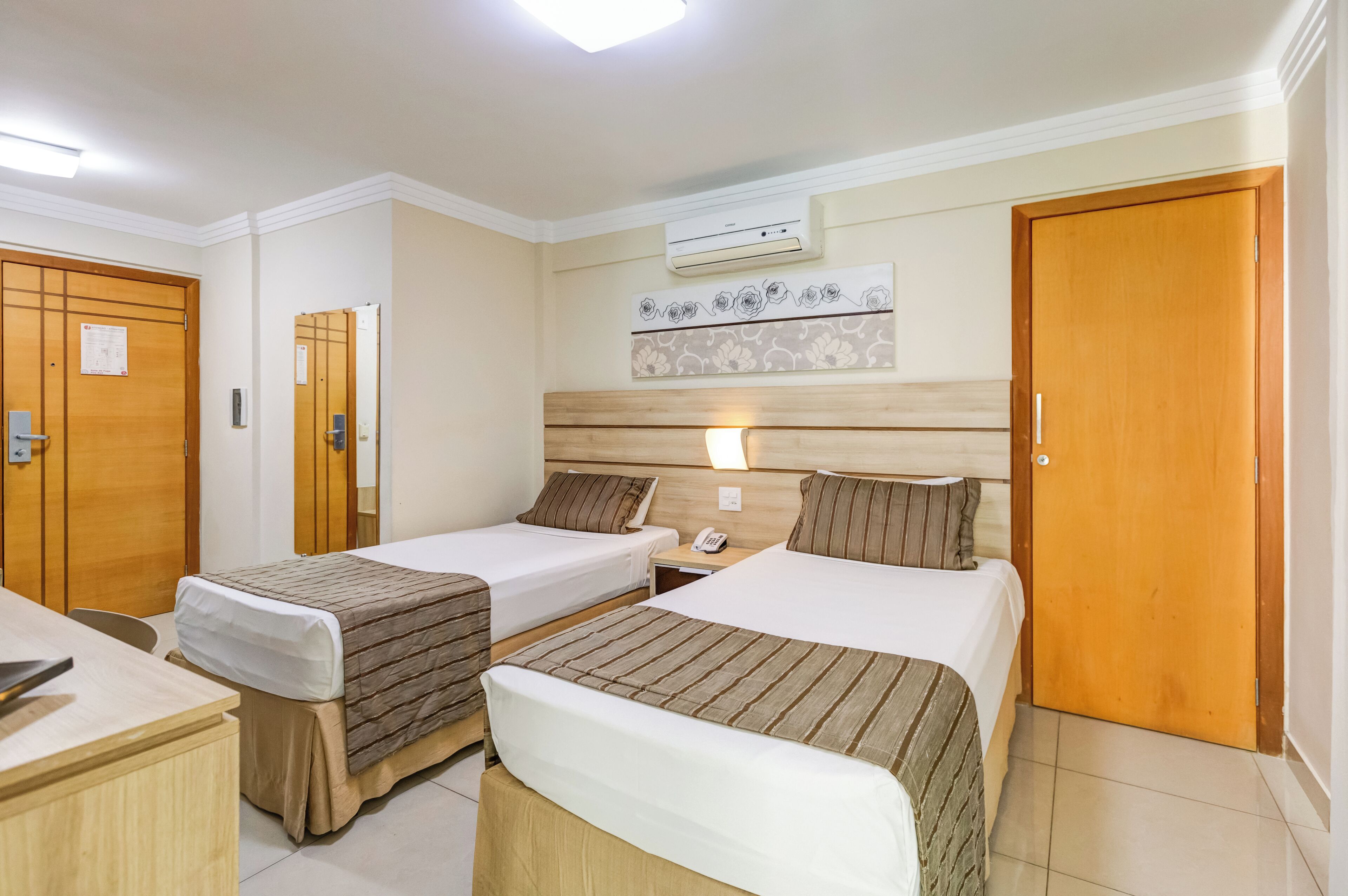 A hotel room with two single beds, a wooden desk, a chair, a bedside table, an air conditioning unit, and a door.