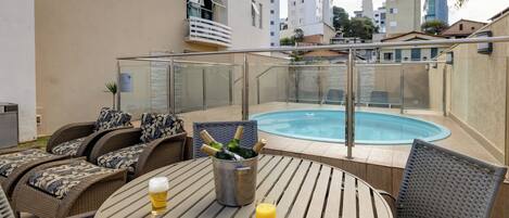 An outdoor seating area with wicker furniture, a glass table, a drink, and a pool.