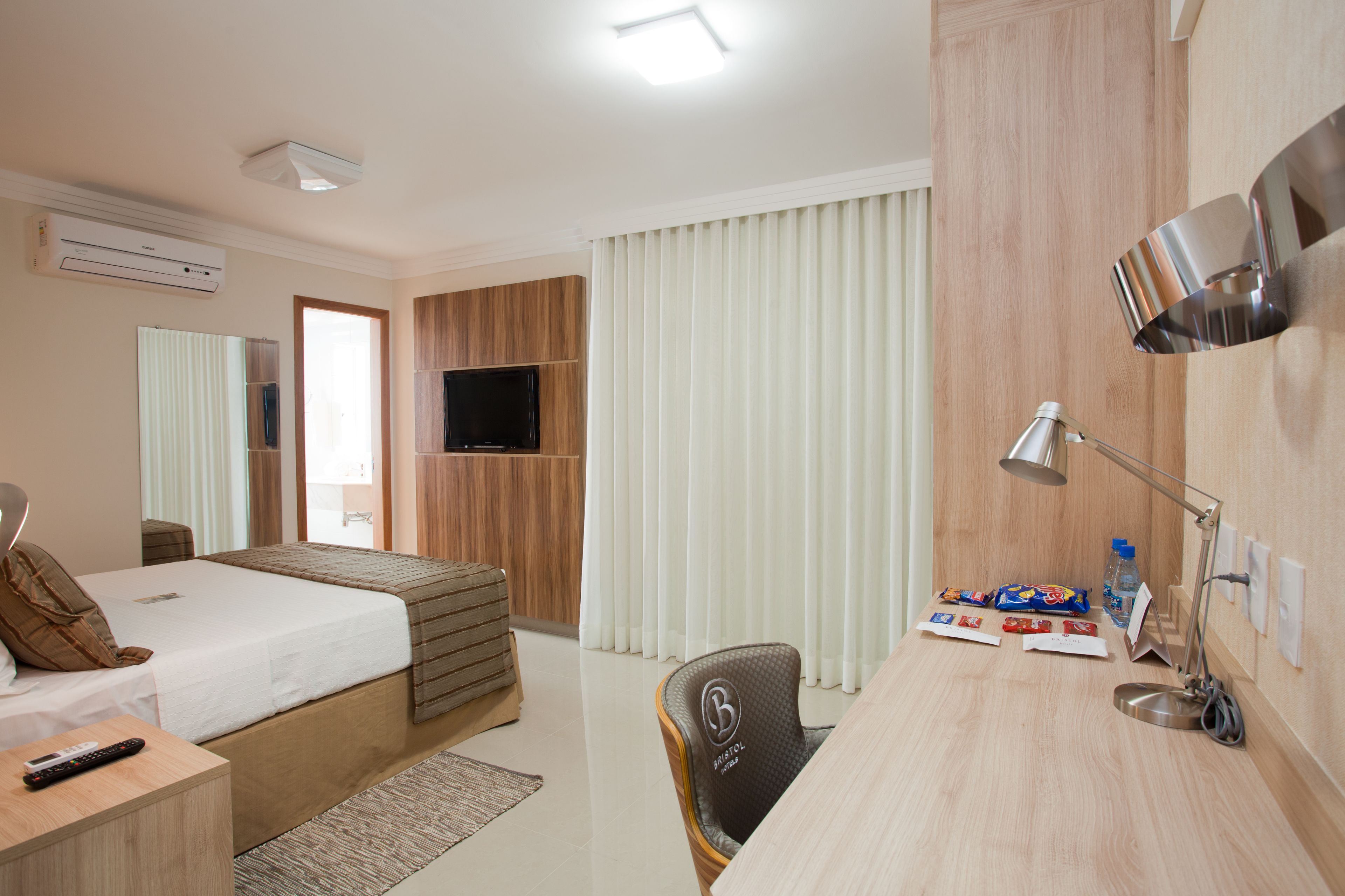 A hotel room with a bed, desk, chair, TV, and a desk lamp.