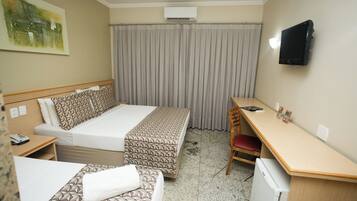 A hotel room with a bed, a desk, a chair, a small refrigerator, an air conditioning unit, and a television mounted on the wall.