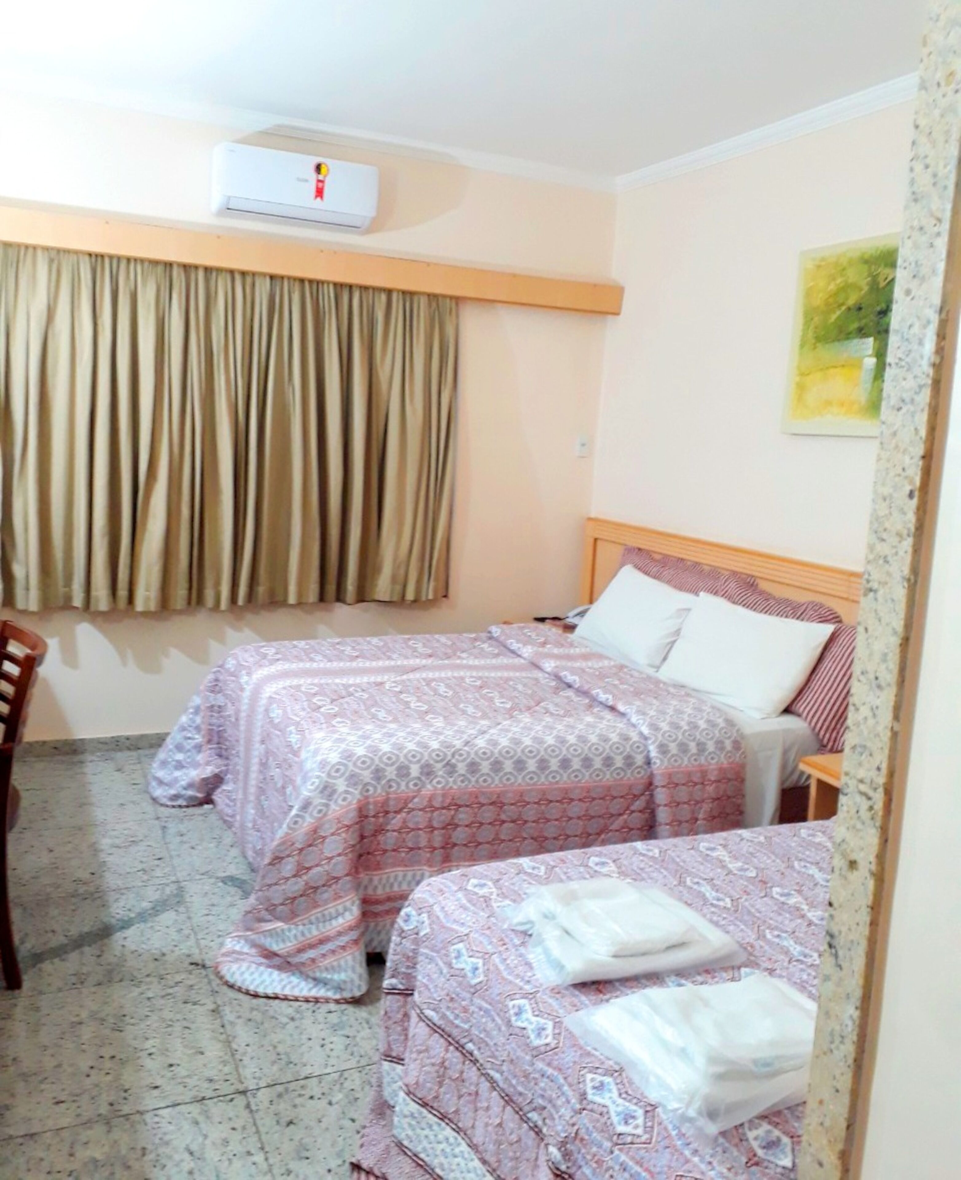 Habitació d'hotel amb dos llits, un tabac de fusta, una finestra amb cortines i un aparell d'aire condicionat a la paret.