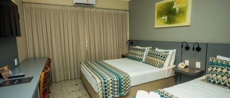 A hotel room with two beds, a desk, a chair, an air conditioner, a painting on the wall, and a telephone on the nightstand.