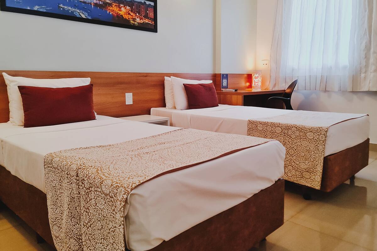 A hotel room with two beds, a desk, a chair, a nightstand, a lamp, and a cityscape painting on the wall.