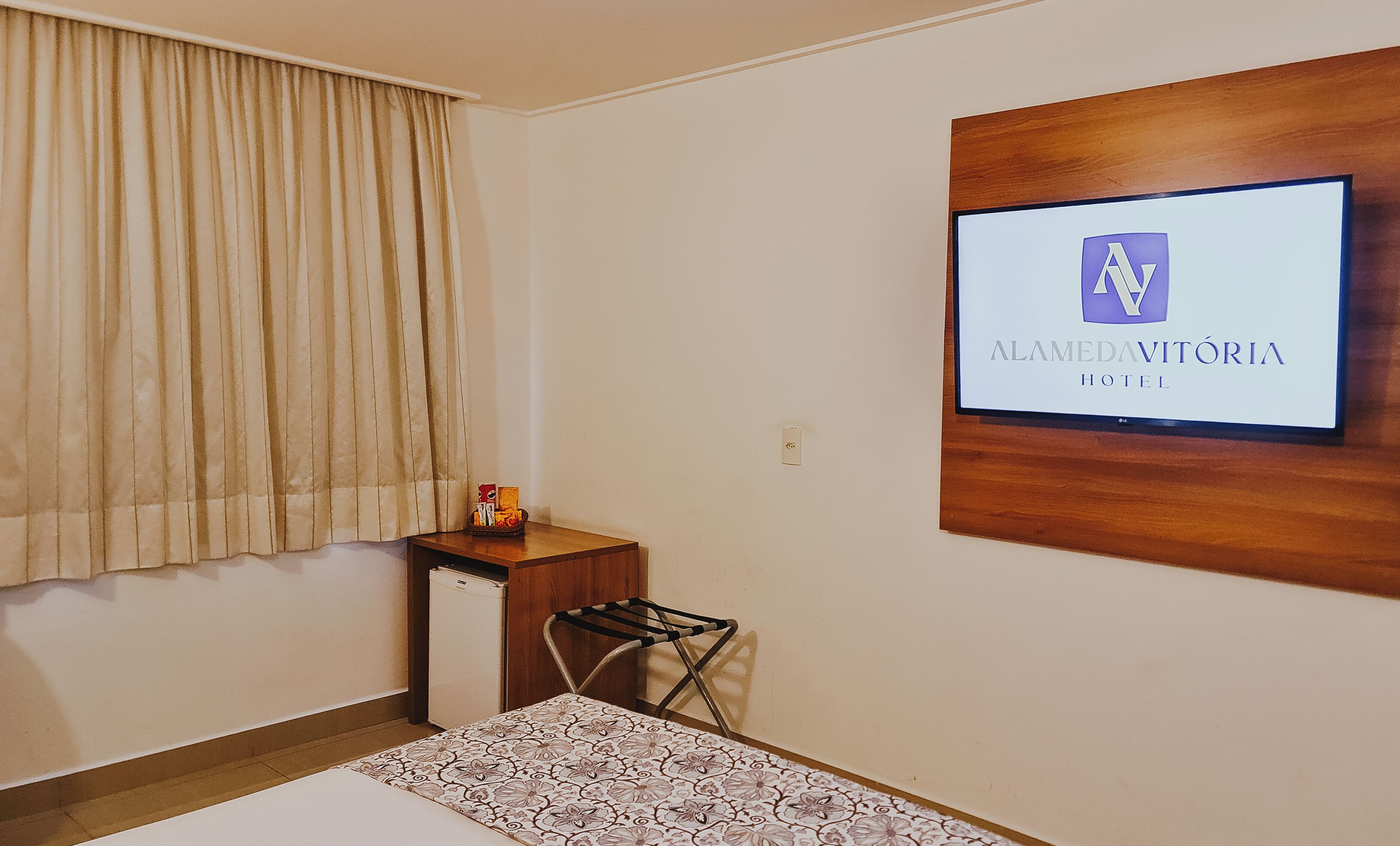 A hotel room with a bed, a small table with a basket, a wall-mounted TV displaying the hotel name, and a window with curtains.
