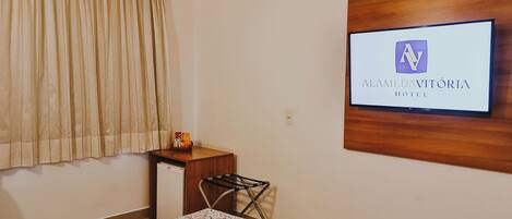 A hotel room with a bed, a small table with a basket, a wall-mounted TV displaying the hotel name, and a window with curtains.
