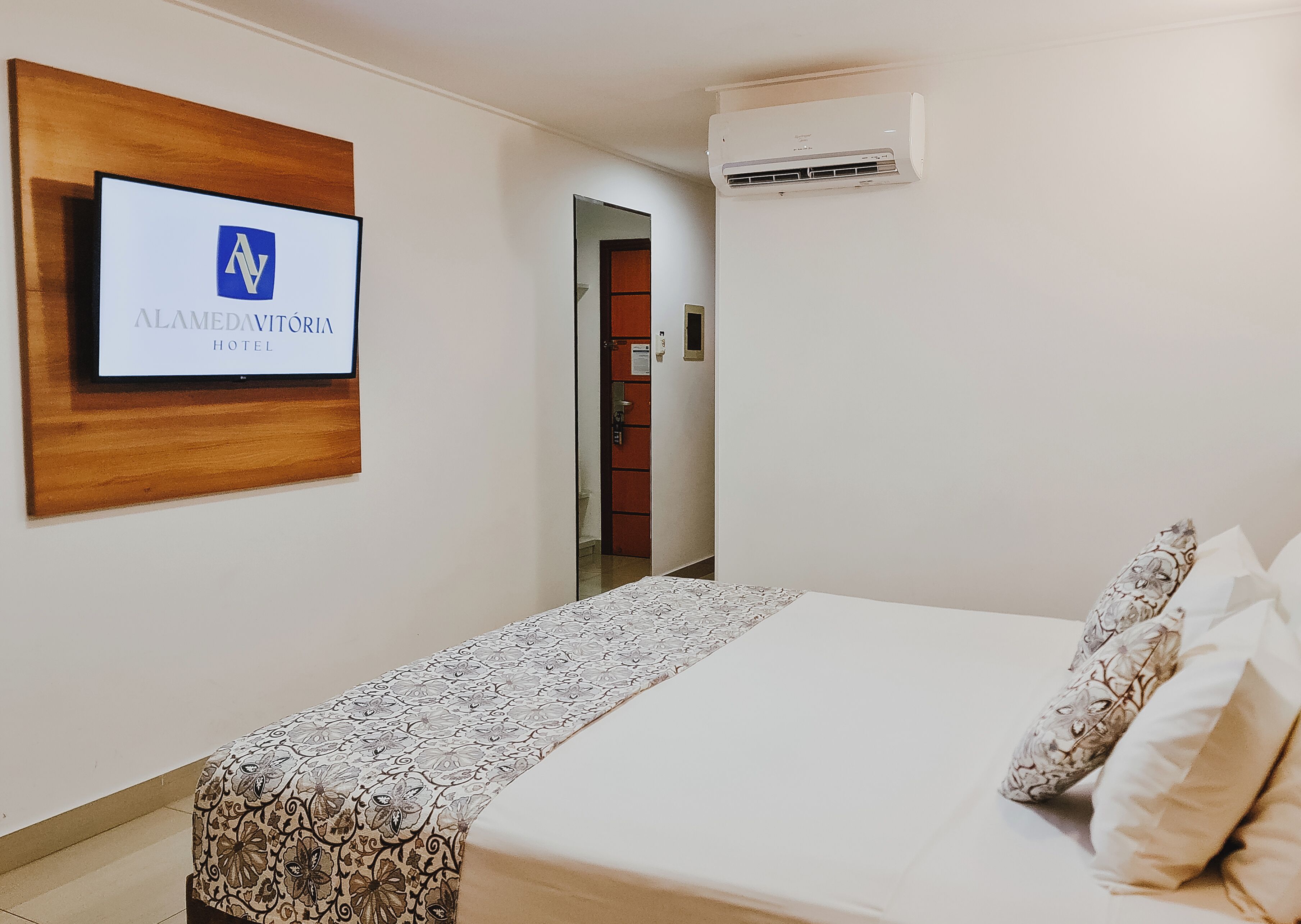 A hotel room with a bed, a TV mounted on the wall, and an air conditioning unit.