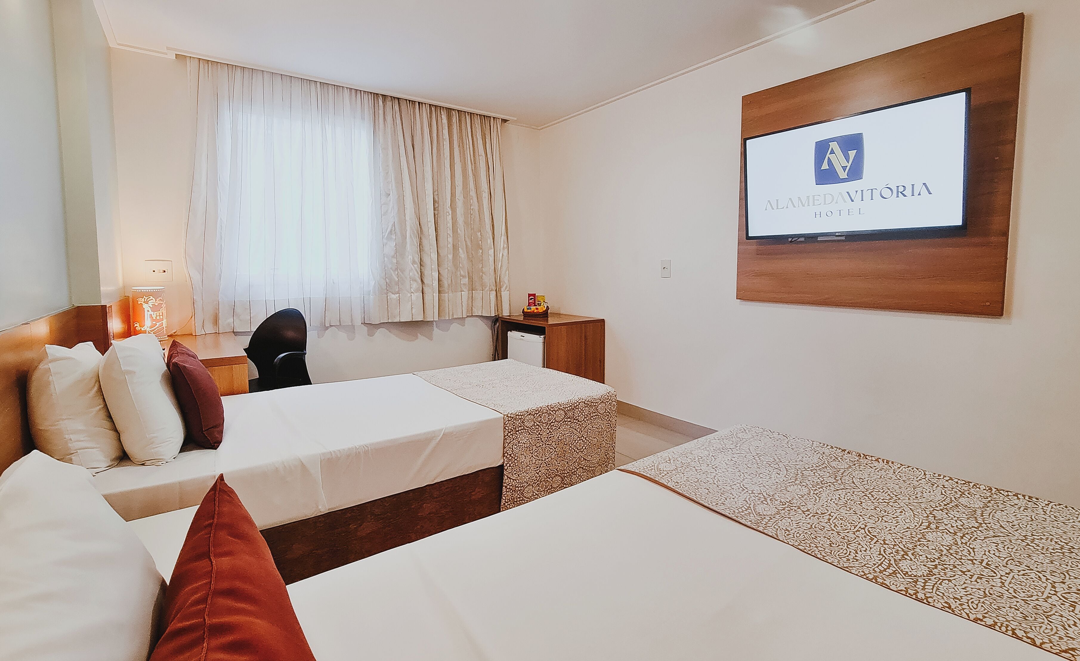 A hotel room with two beds, a desk, a chair, a TV mounted on the wall, and a window with curtains.