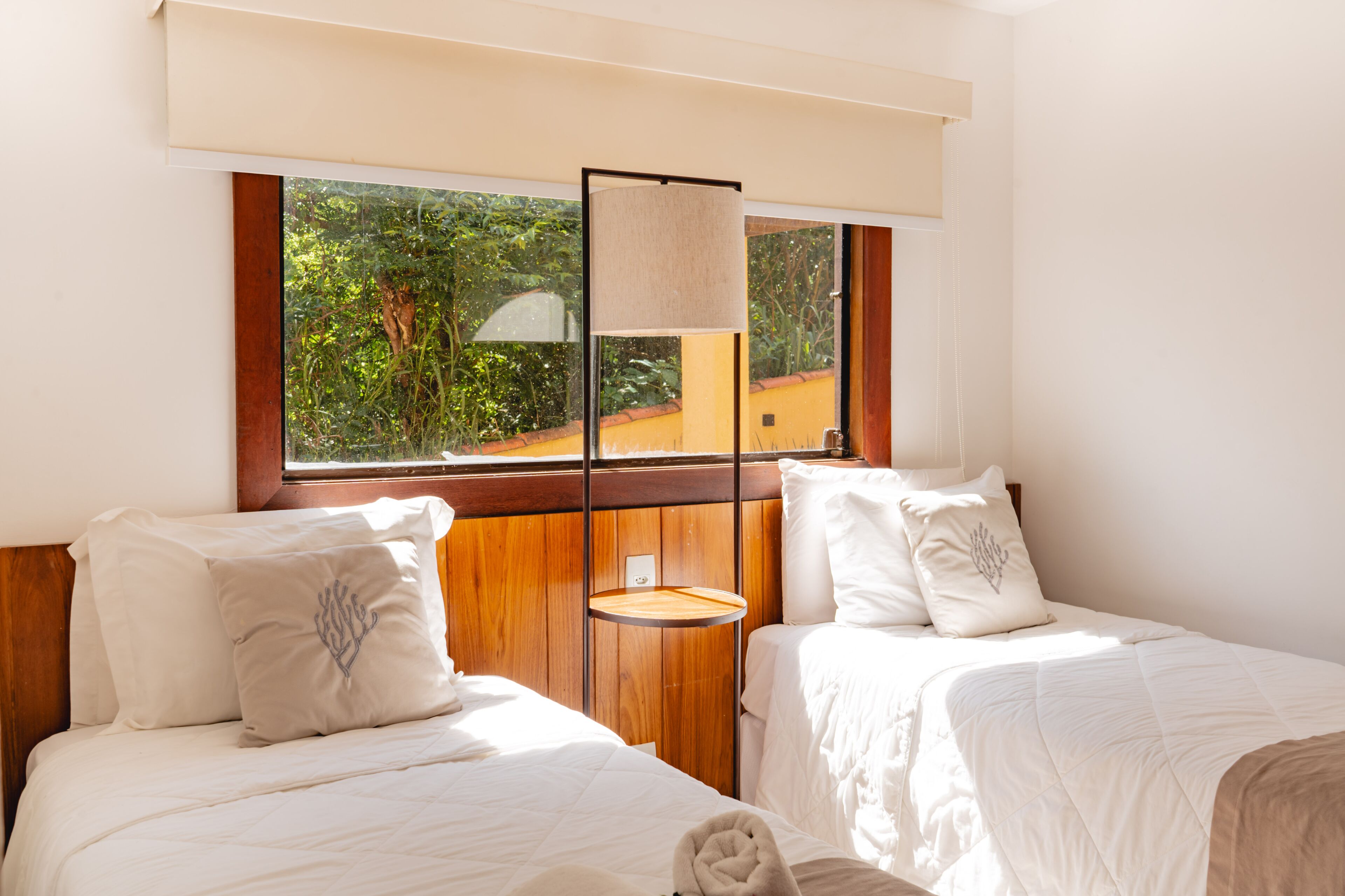 Une chambre d’hôtel avec deux lits, une fenêtre donnant sur un paysage verdoyant, une tête de lit en bois et une table de chevet.