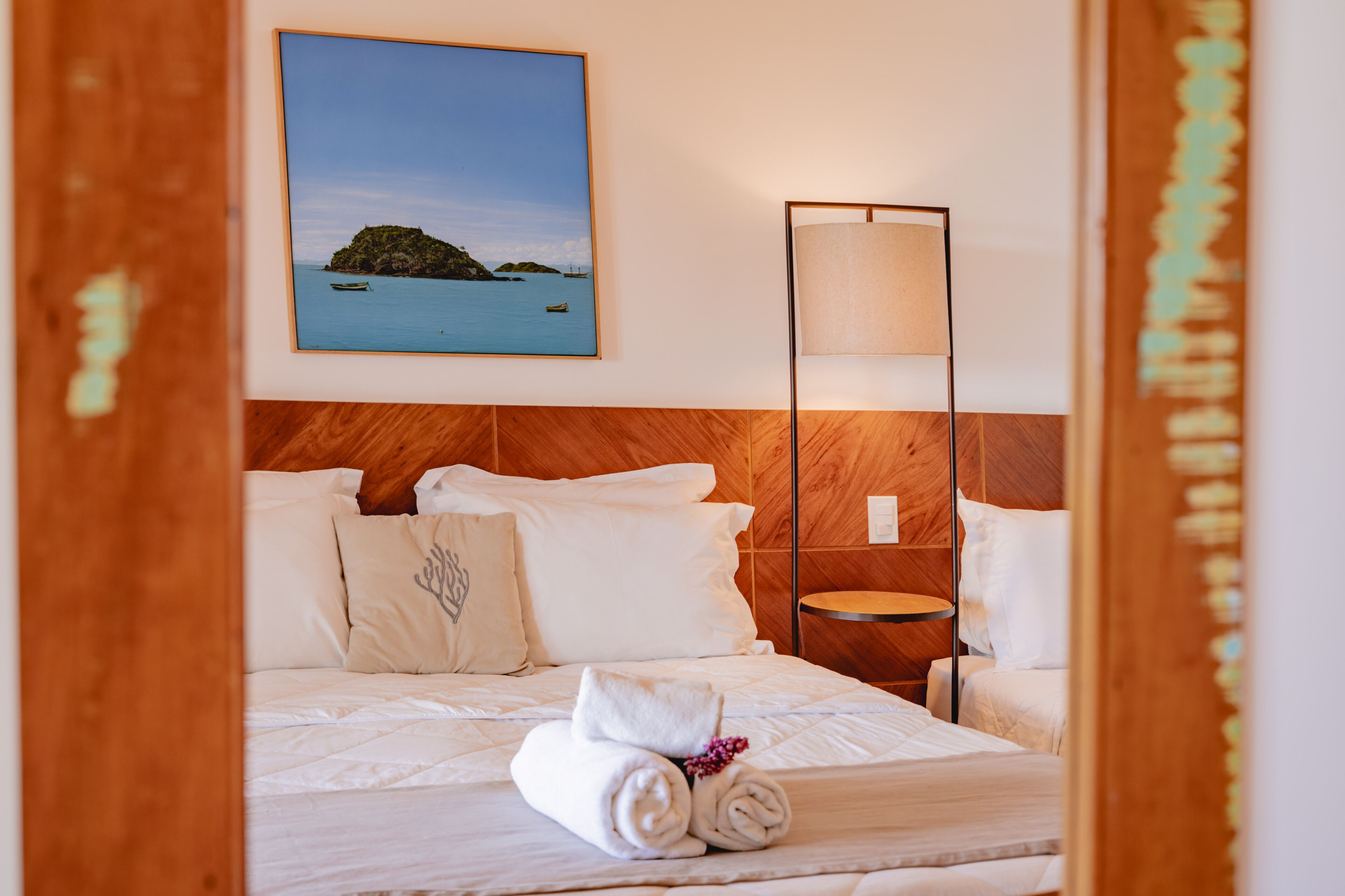 A hotel room with a bed, pillows, a rolled towel, a nightstand, a lamp, and a framed picture of a coastal scene.