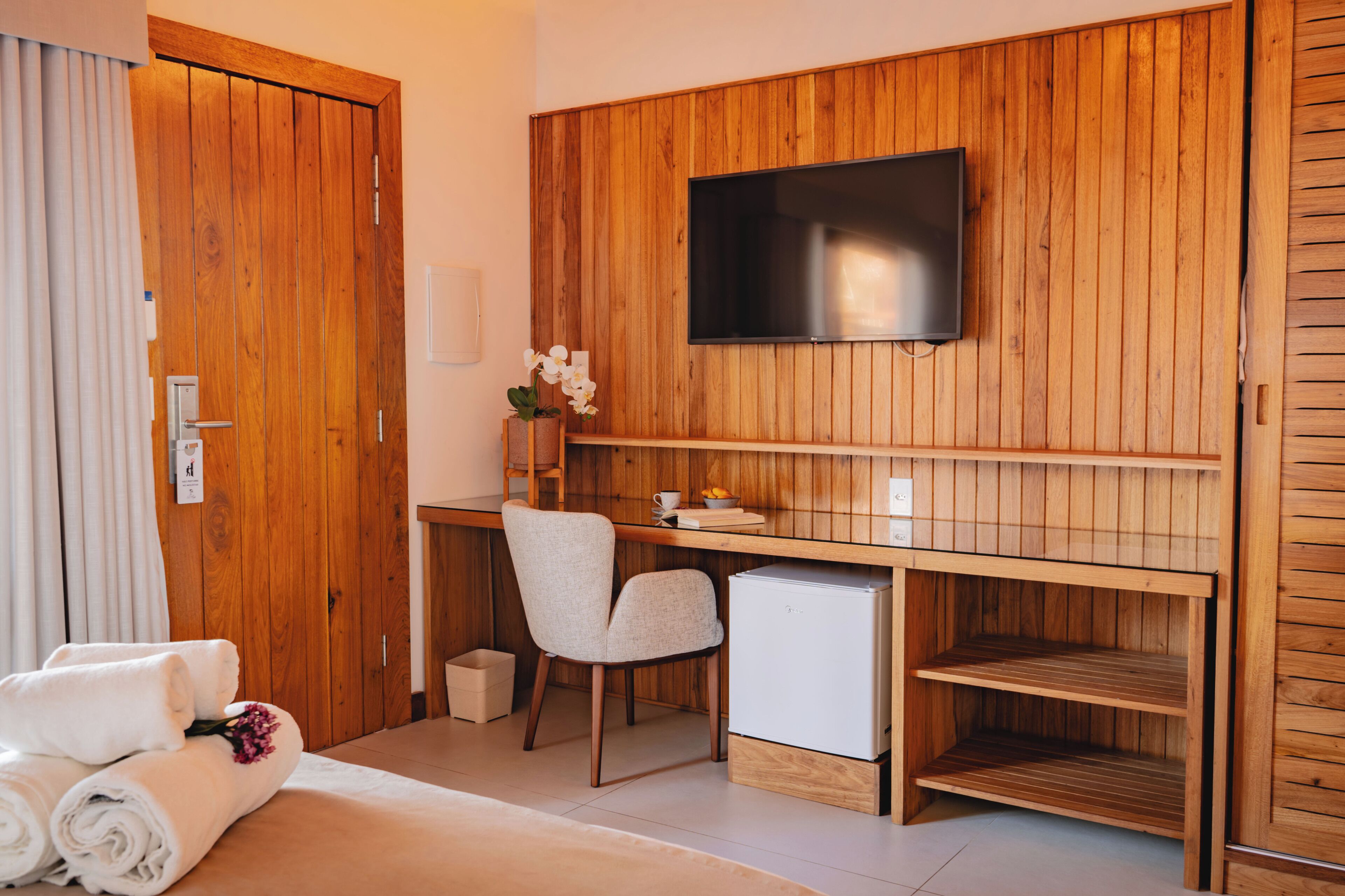 Ein Hotelzimmer mit einem Holztisch, einem Fernseher, einem Sessel, einer kleinen Kühlbox und einem Bett mit zusammengerollten Handtüchern.