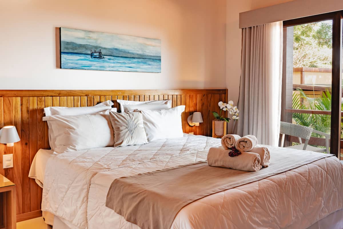 A neatly made bed with a wooden headboard, a painting of a boat on the wall, and a view of greenery through the window.