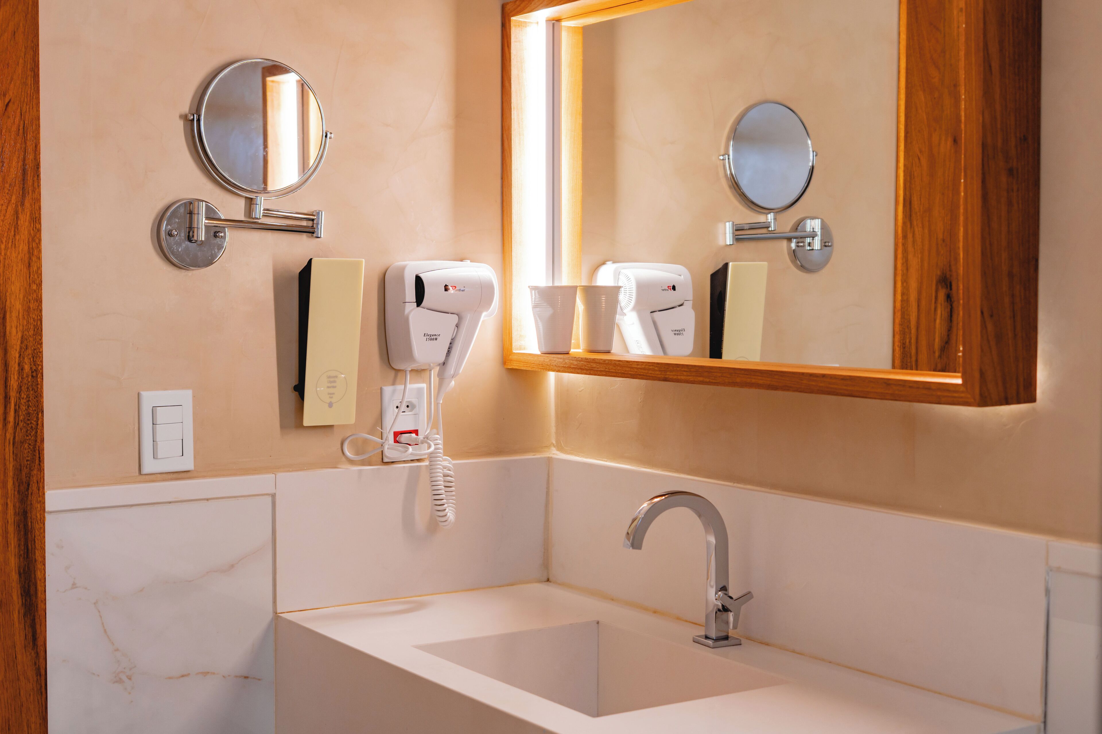 Un baño con lavamanos, dos espejos, secador de pelo, portavasos y un interruptor de luz.