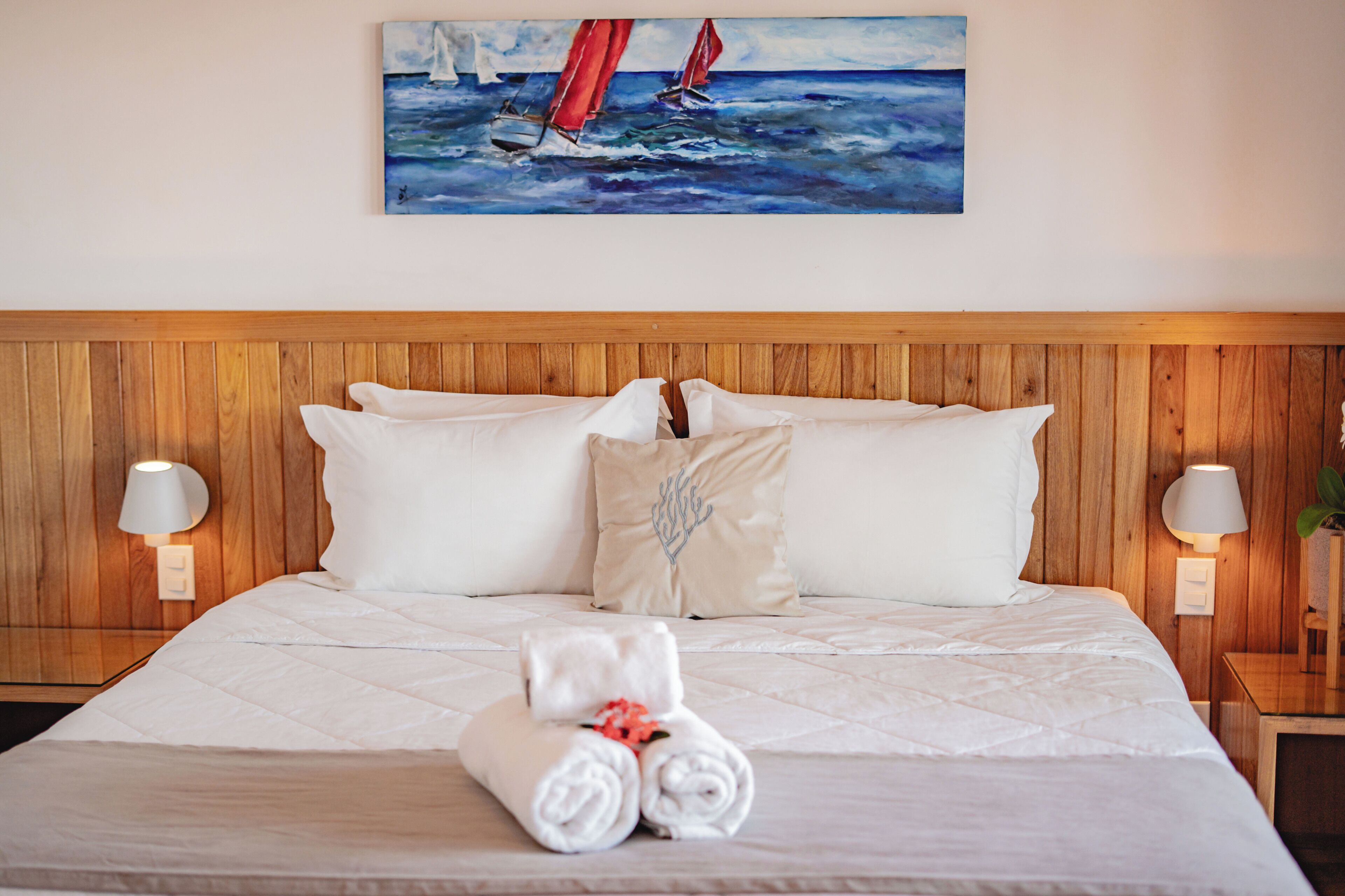Un lit bien fait, avec du linge de lit blanc, un tableau représentant des voiliers accroché au mur et un panneau de bois ornant la tête de lit.