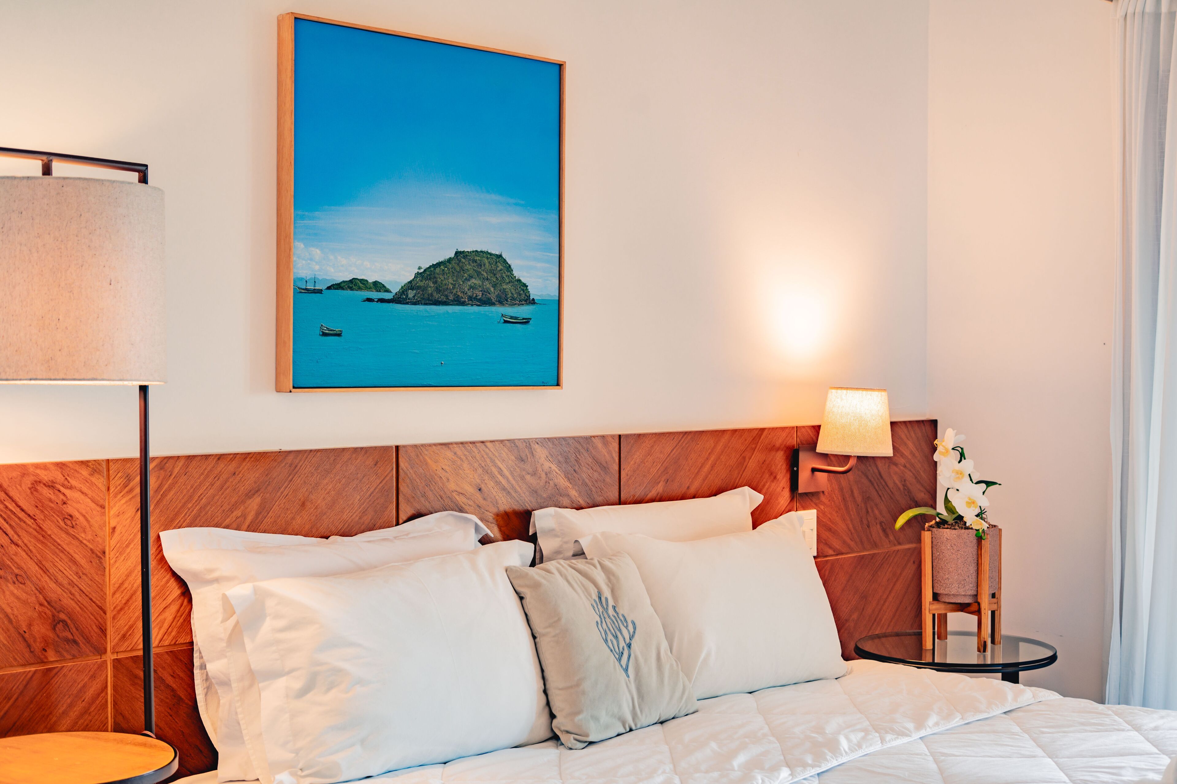 A hotel room with a wooden headboard, a bed with white bedding, a round bedside table with a lamp, and a framed picture of a coastal scene.