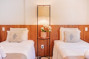 Due letti con biancheria da letto bianca e cuscini coordinati, un piccolo comodino in legno con un'orchidea in vaso tra di essi, e una lampada fissata al muro.
