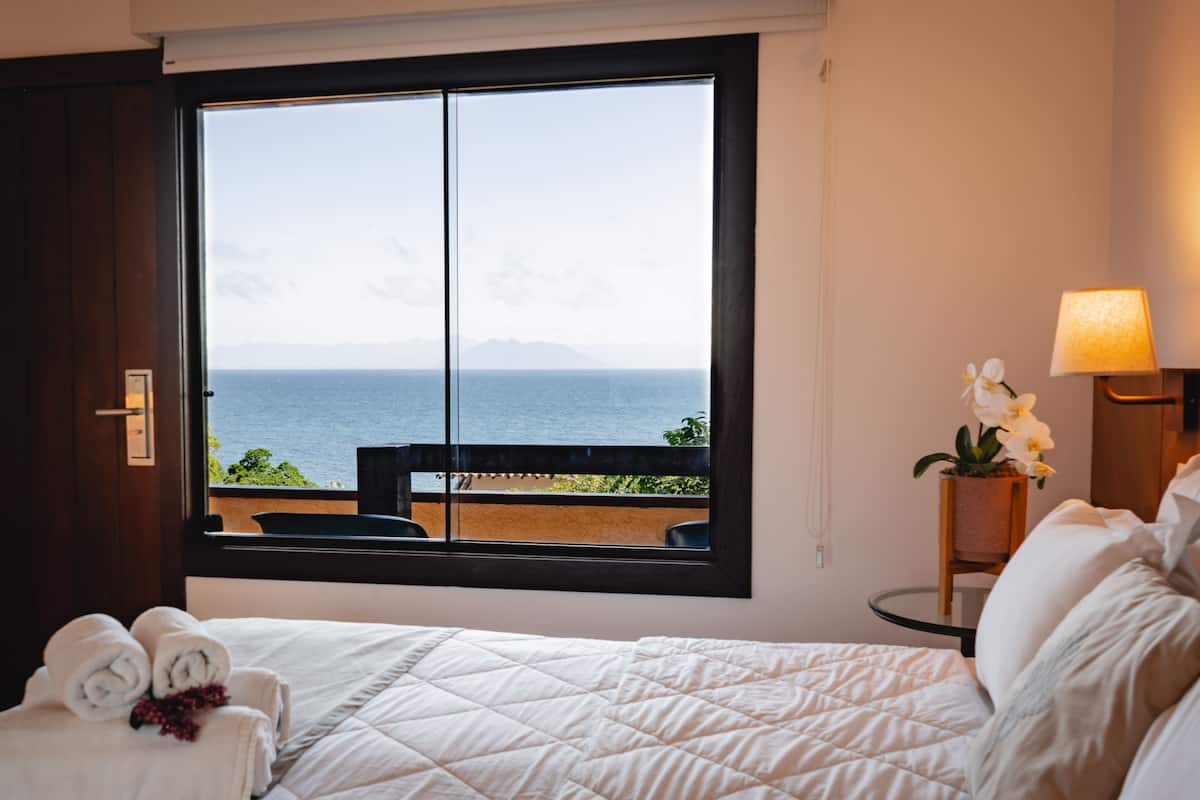 A hotel room with a bed, a window with a view of the sea, a small table with a lamp, and a potted orchid.
