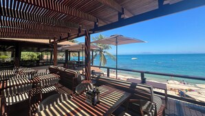 Un espace extérieur aménagé avec des tables et des chaises en bois, des parasols et une vue sur l’océan.