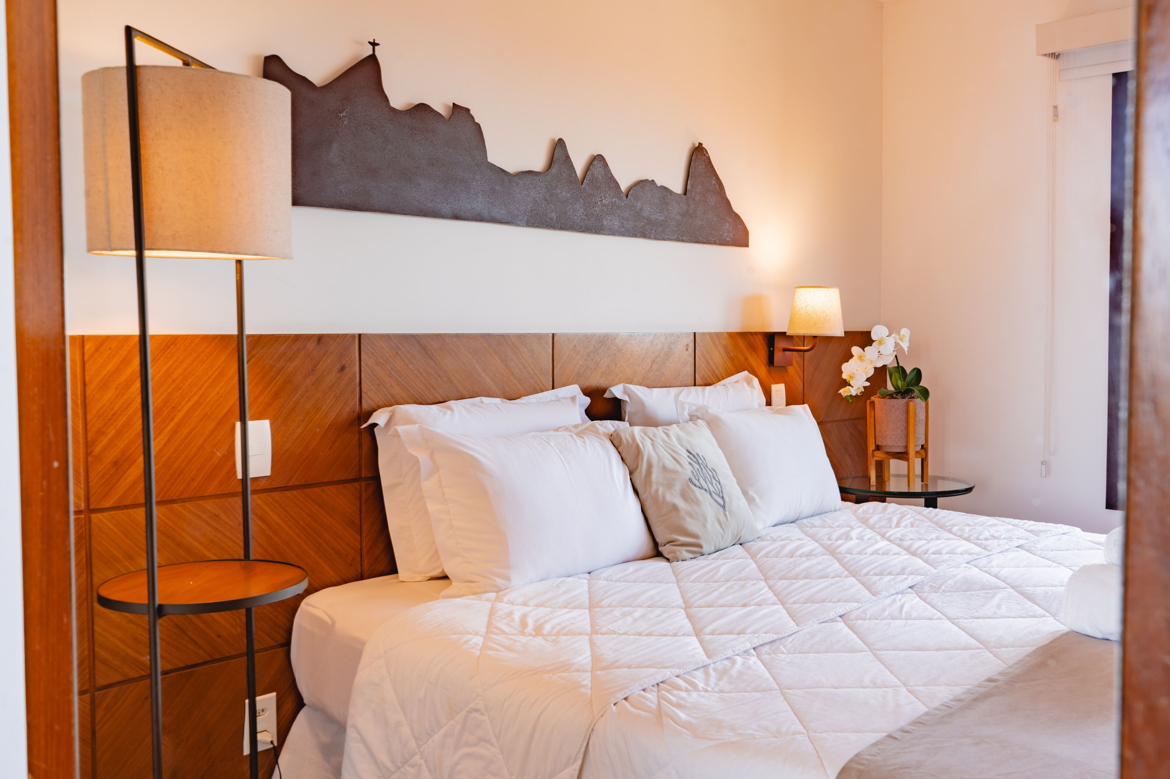 Una cama bien hecha con ropa blanca, un cabecero de madera, una lámpara de pared y una mesita con un florero.