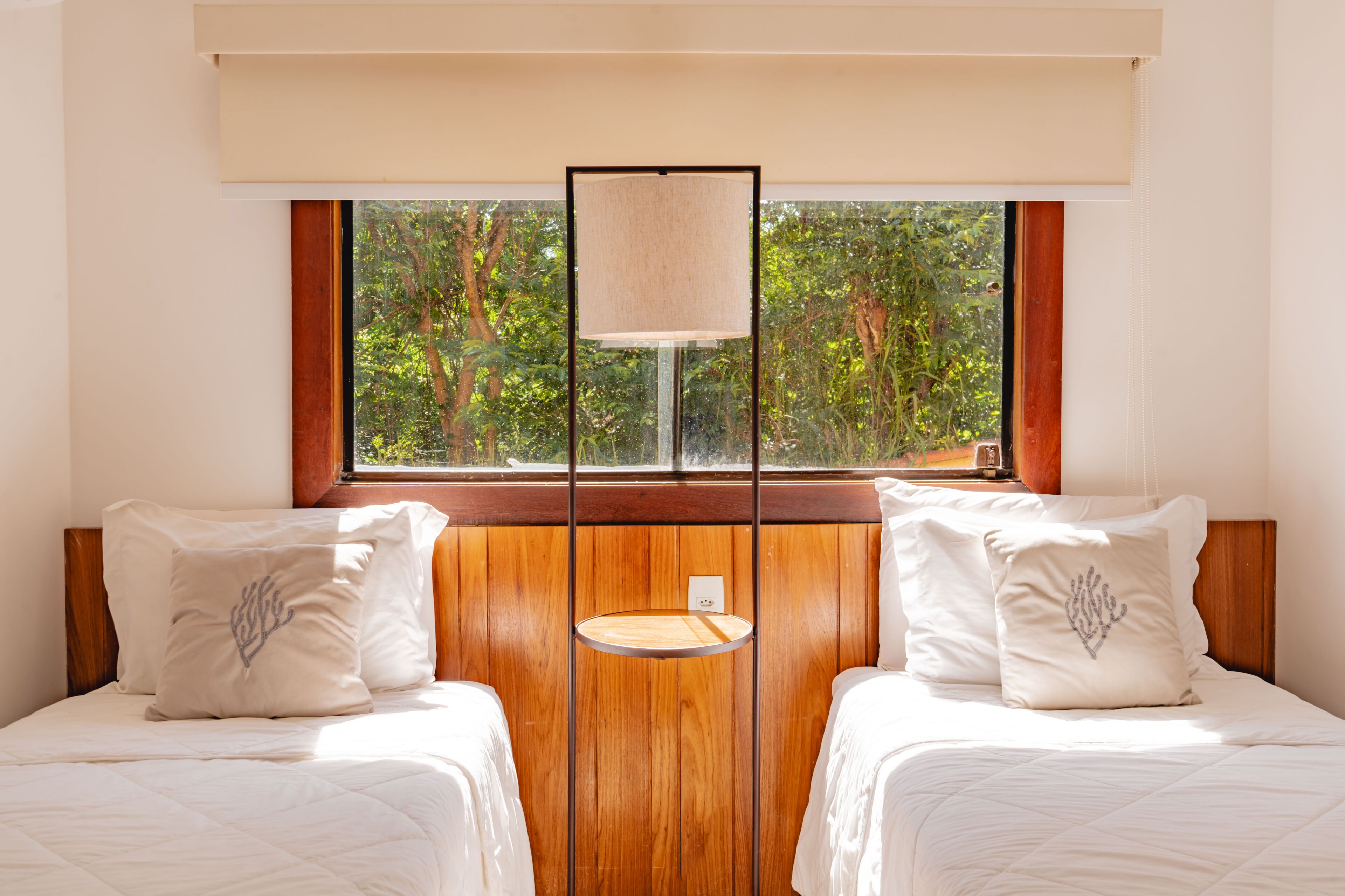 A room with two beds, a window with a view of trees, and a round table with a lamp.