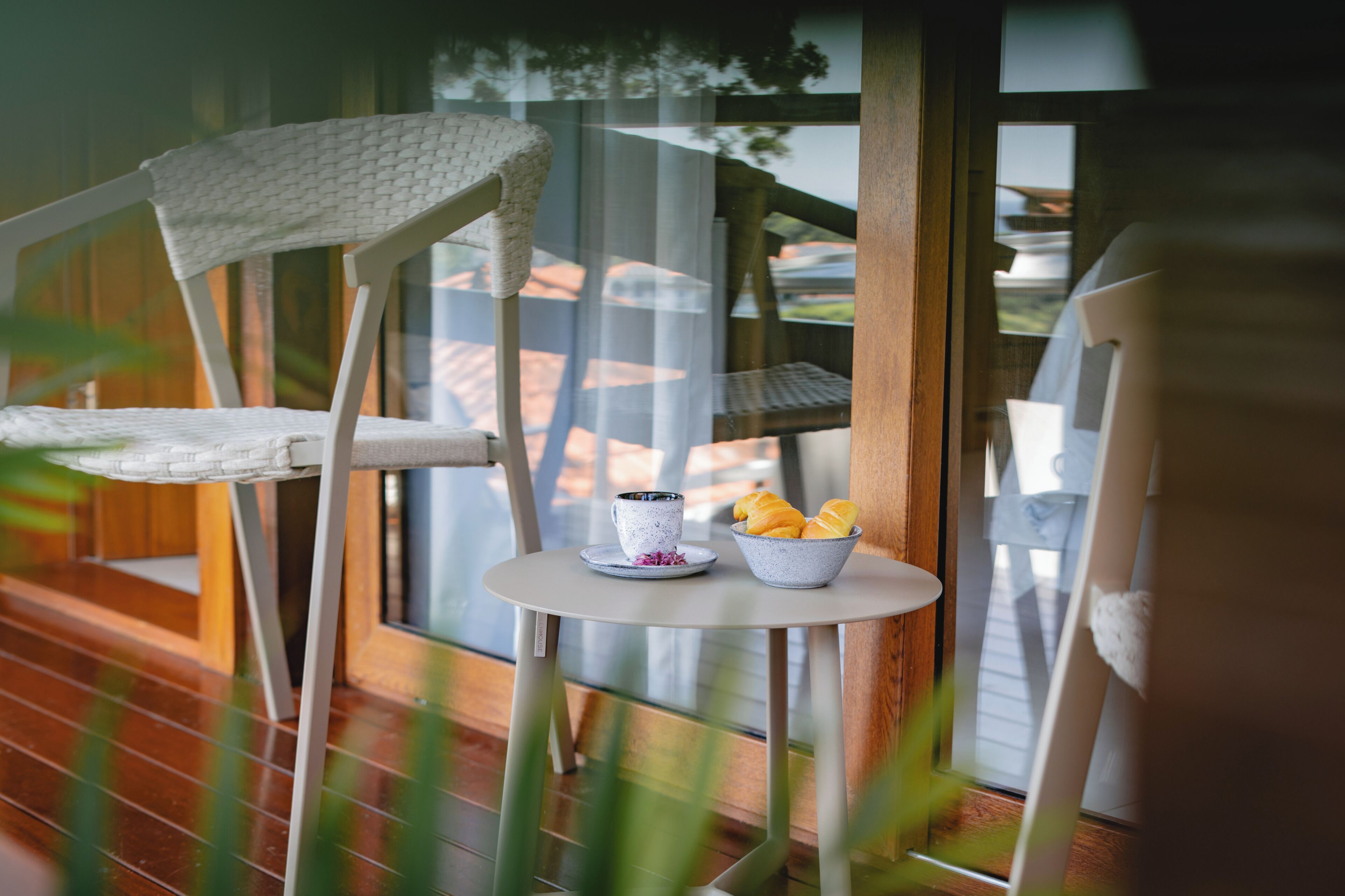 Une chaise blanche avec un dossier en osier, une petite table ronde sur laquelle se trouve un bol blanc contenant des croissants, et une tasse posée sur la table.