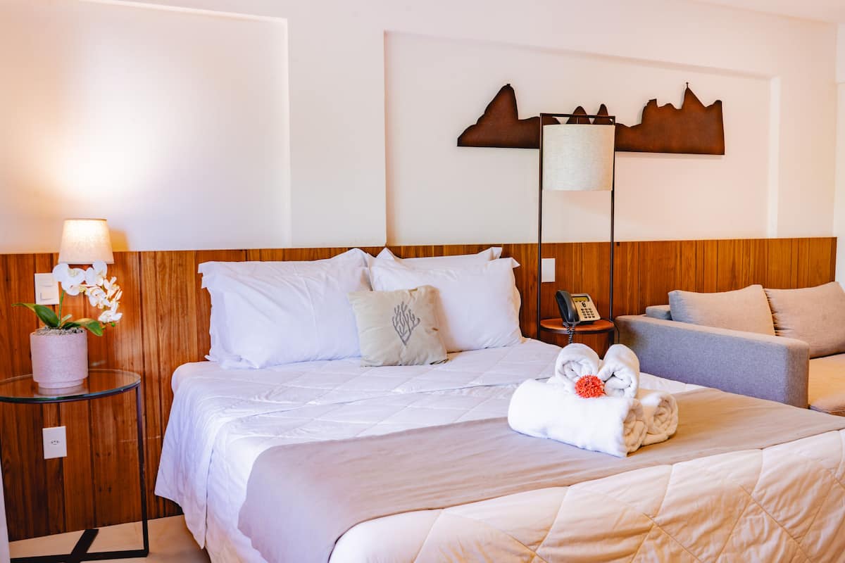 A hotel room with a bed, a bedside table with a lamp, a small potted plant, and a wall-mounted artwork.