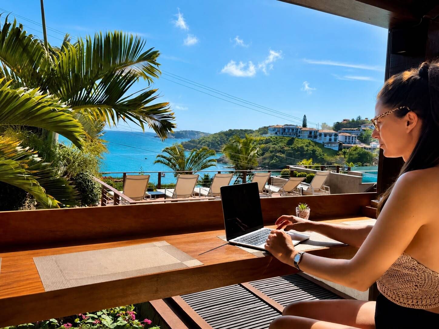 A person working on a laptop at a wooden table with a view of the sea and buildings in the background.