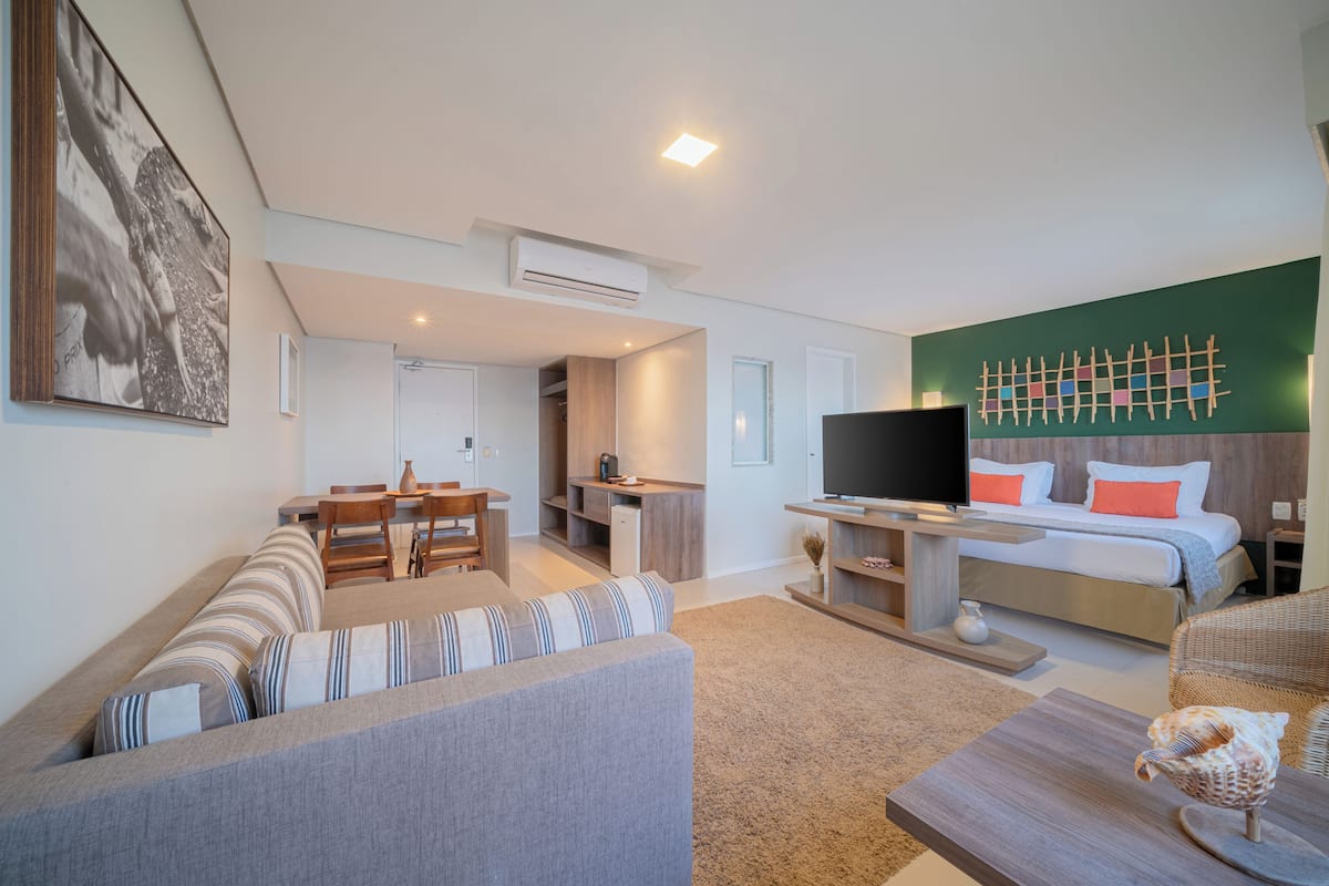 A modern living room with a sofa, a dining area, a TV, and a bedroom with a bed and a wall art piece.