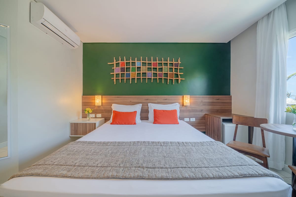 A hotel room with a large bed, two bedside tables with lamps, a wooden headboard, a green accent wall, and a view of the outdoors.