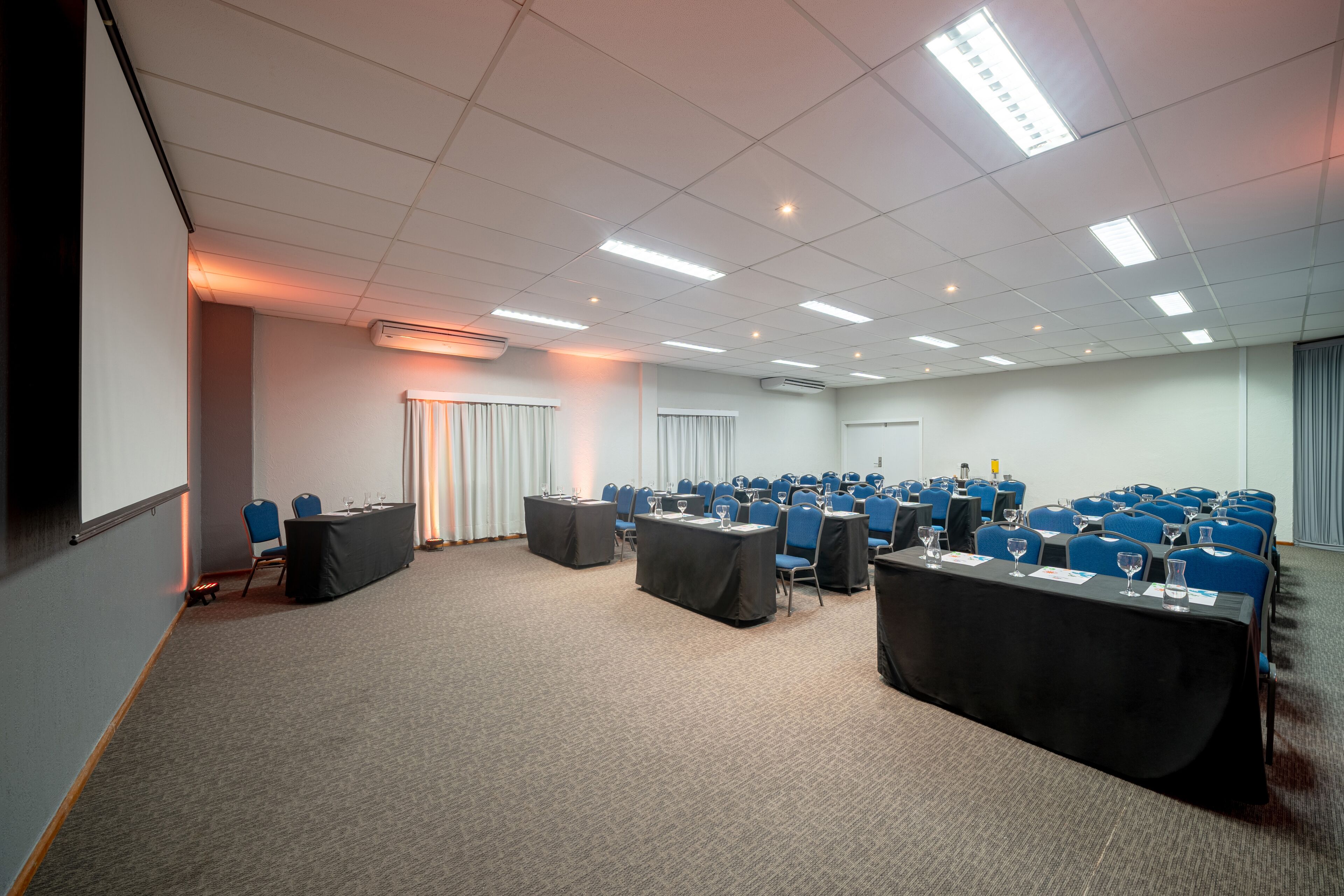 Een vergaderzaal met rijen blauwe stoelen, een projectiescherm en tafels met waterglazen.