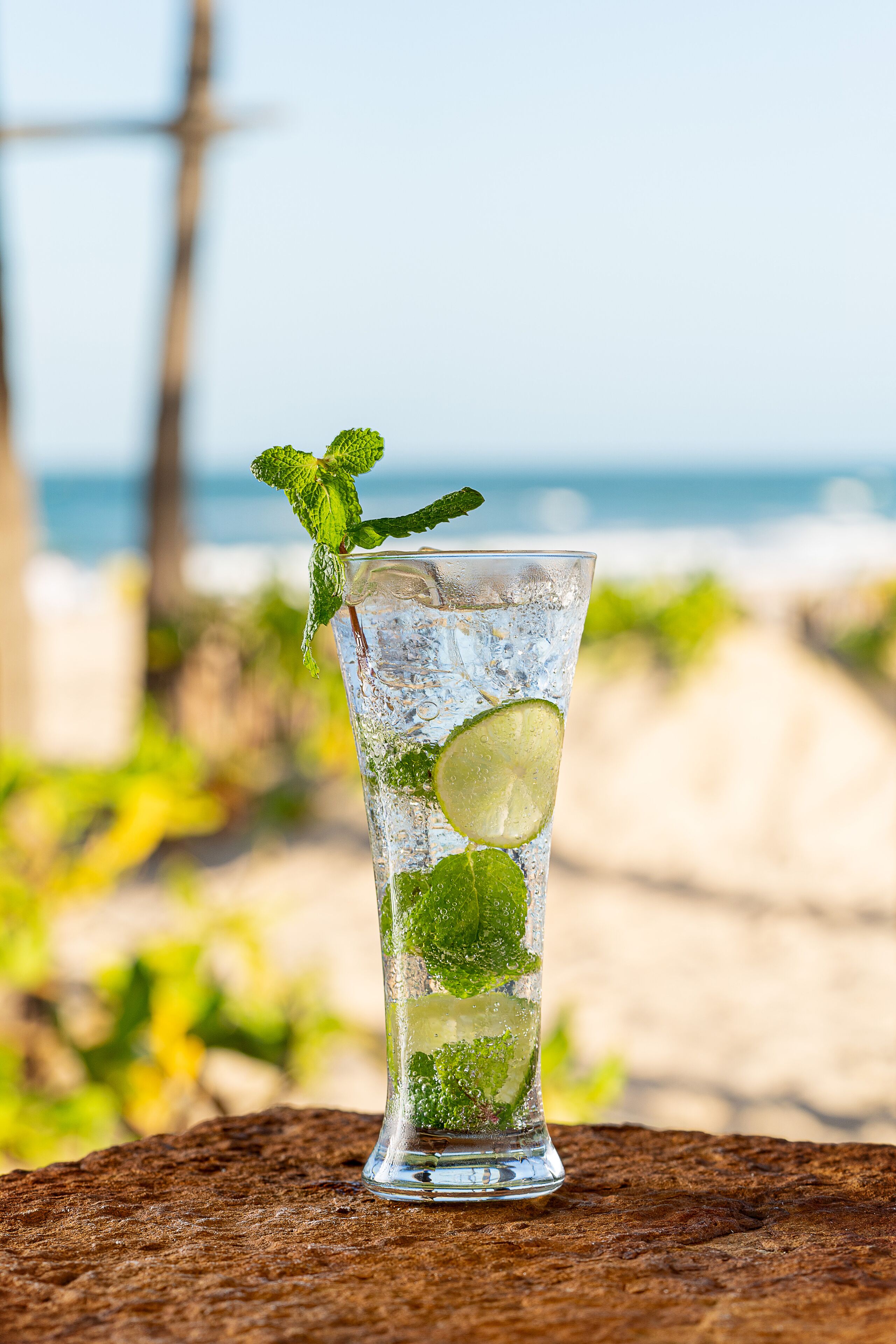 A tall glass of a refreshing beverage with lime and mint, placed on a wooden surface with a beach and ocean in the background.