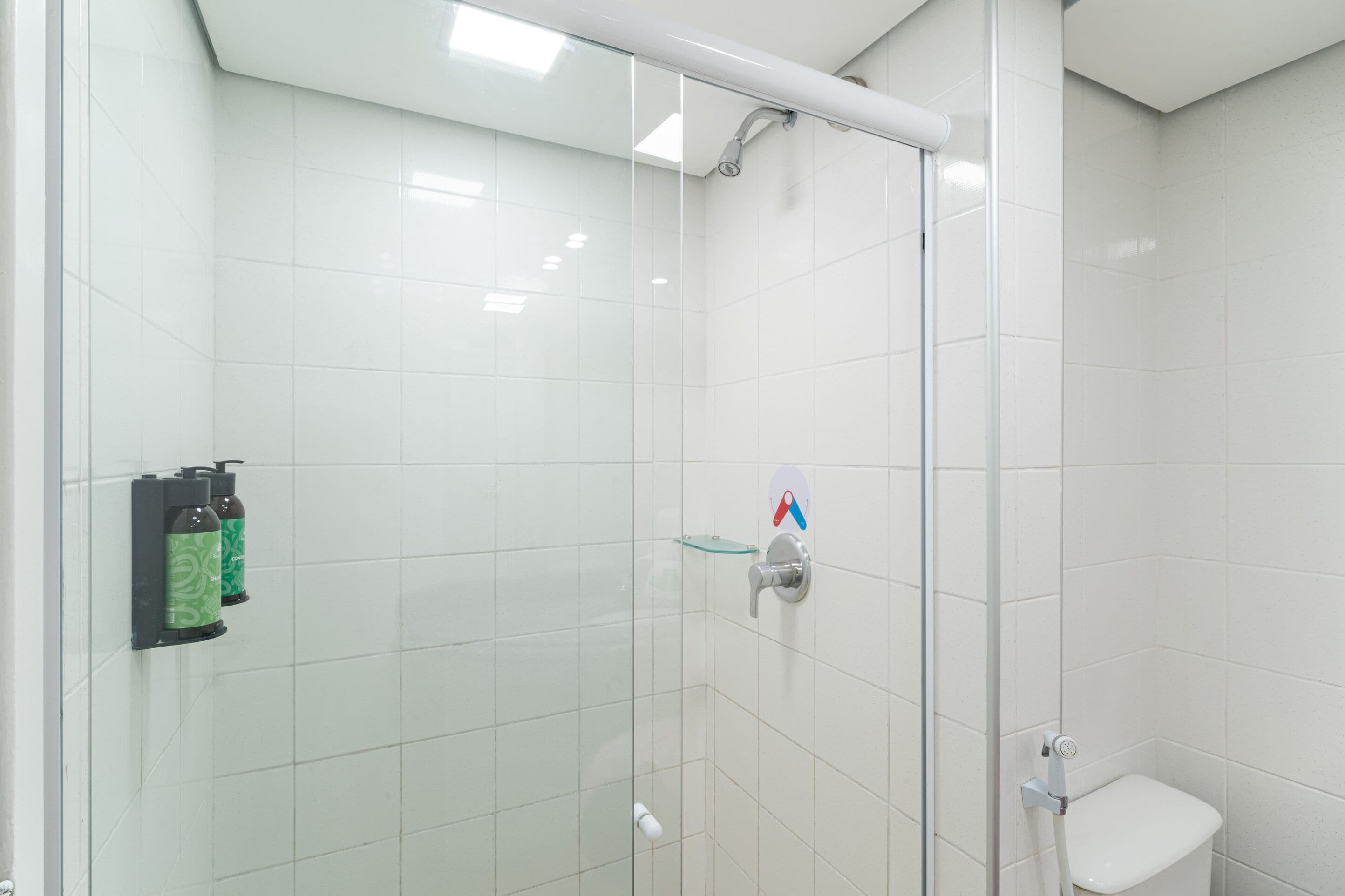A glass-enclosed shower stall with white tiles, a toilet, and two green-patterned soap dispensers on a wall-mounted holder.