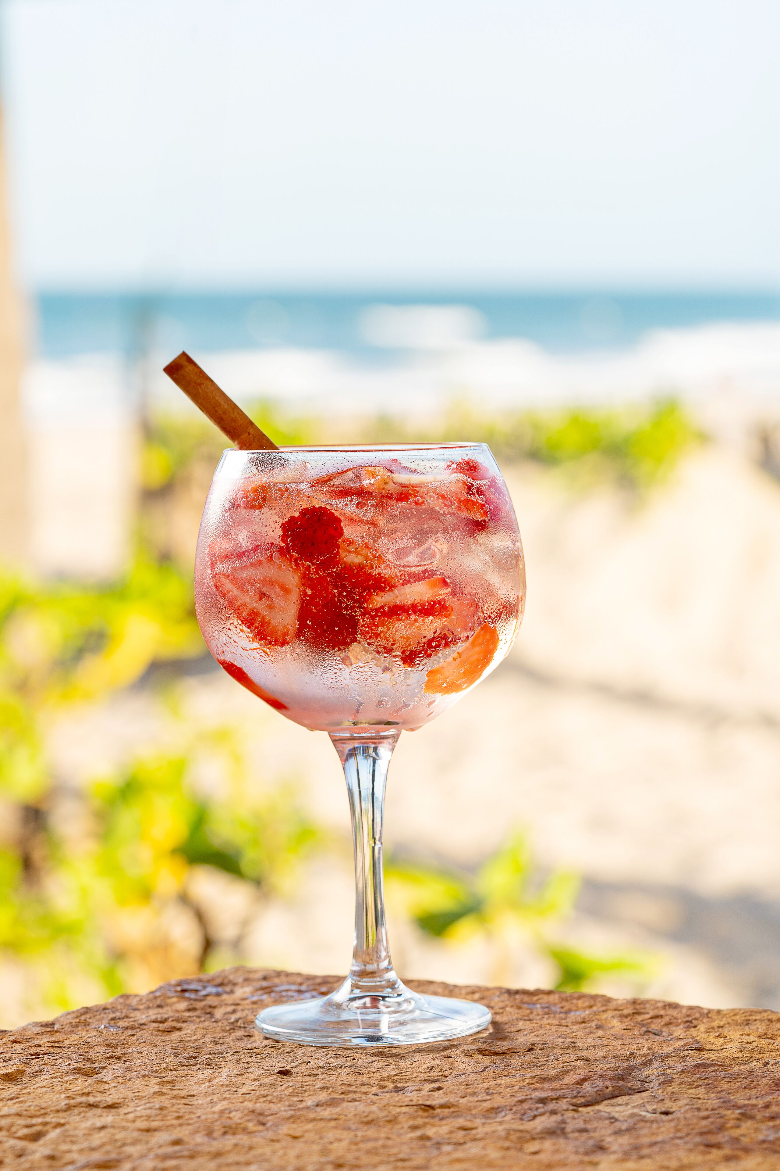 Un vaso de cóctel con una pajita sobre una superficie rugosa, posiblemente una mesa, con una playa y océano desenfocados al fondo.