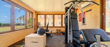 Een sportschool met trainingsapparatuur, een houten doos met de tekst 'STUDIUM FITNESS' en grote ramen met uitzicht op palmbomen.