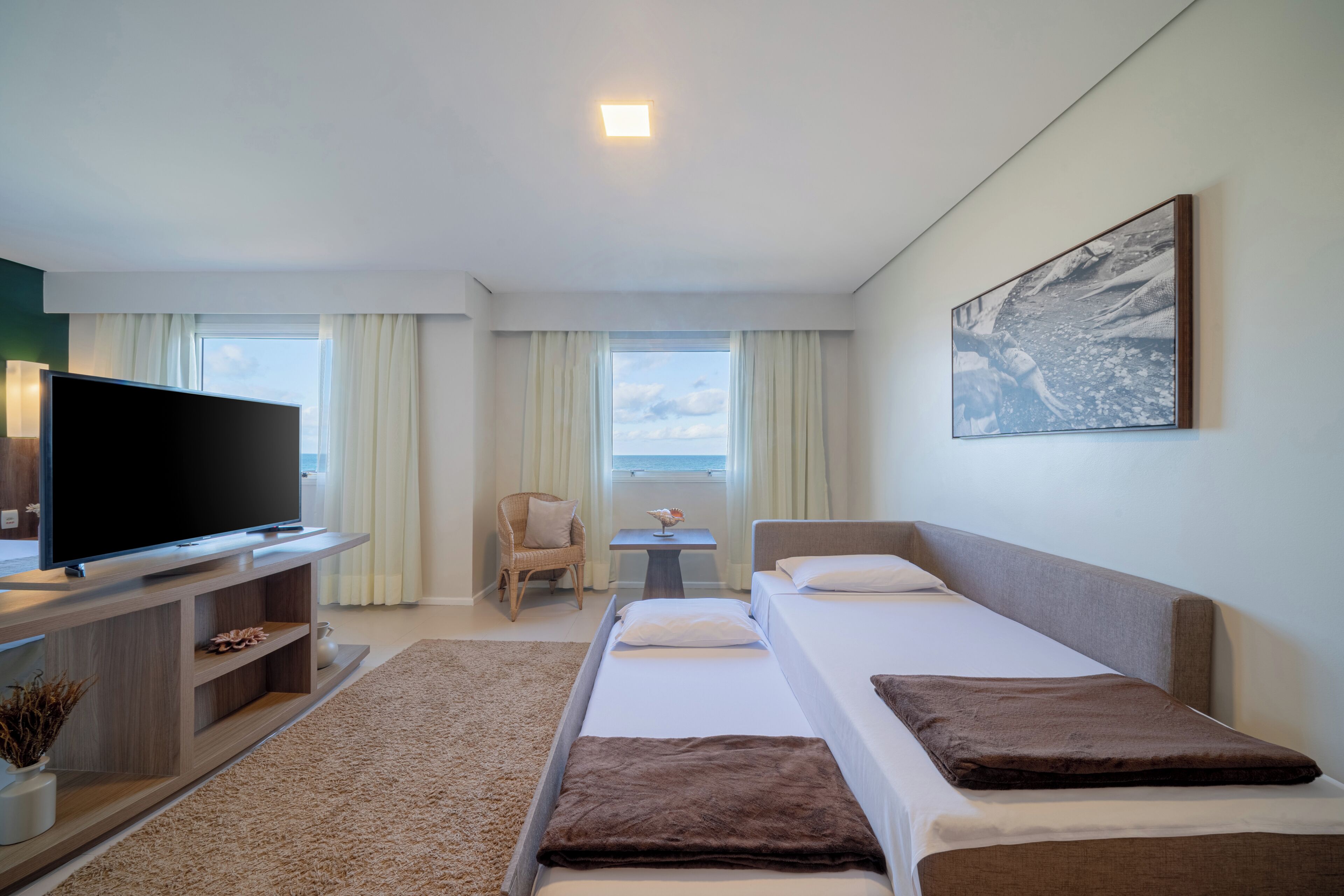 A hotel room with a TV on a wooden stand, two beds with brown pillows, a small table with a vase, and a window with curtains.