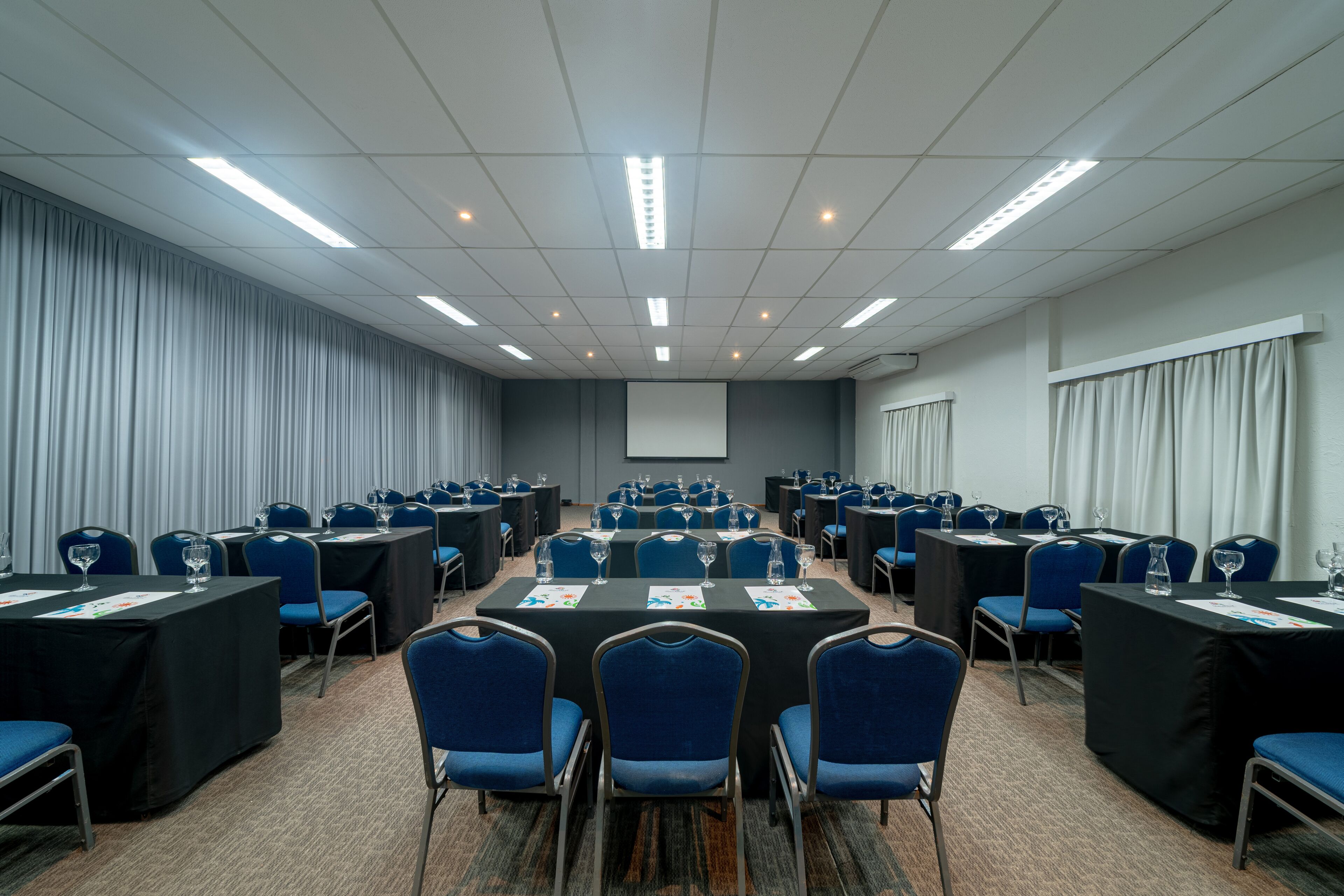Un salón de conferencias con varias mesas y sillas dispuestas en filas, y una pantalla de proyector al frente.