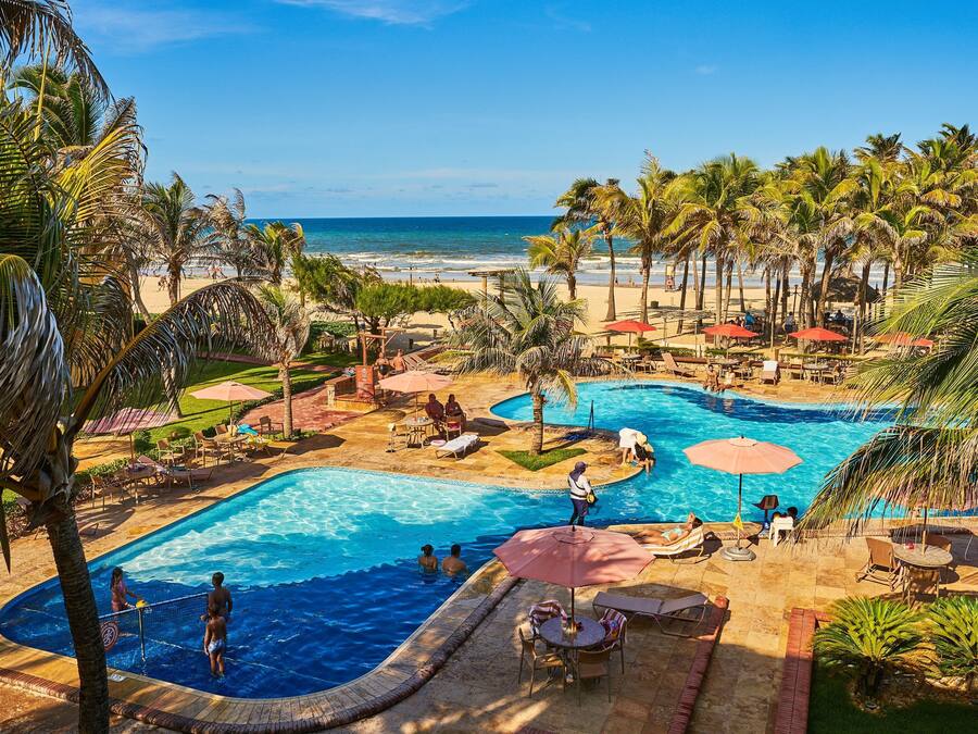 A resort pool area with a view of the beach, palm trees, and sun loungers.